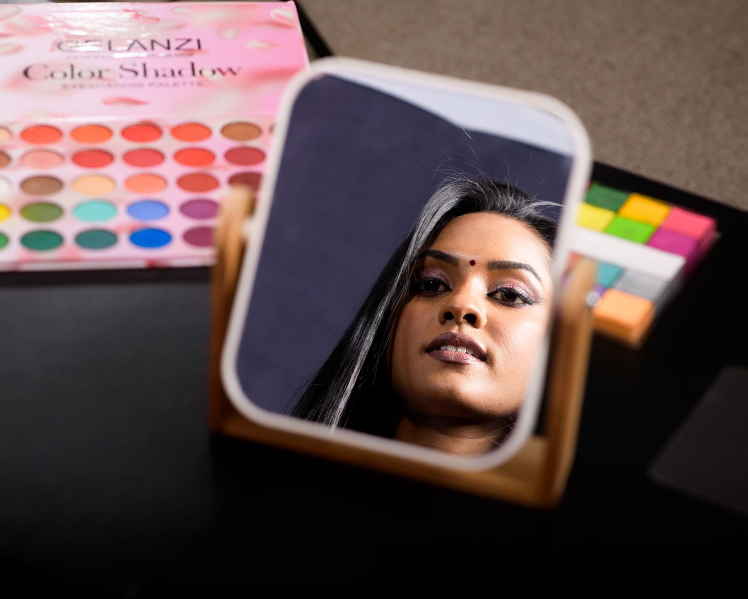Woman applying makeup reflected in mirror beside TANZI Color Shadow eyeshadow palette on dark surface.