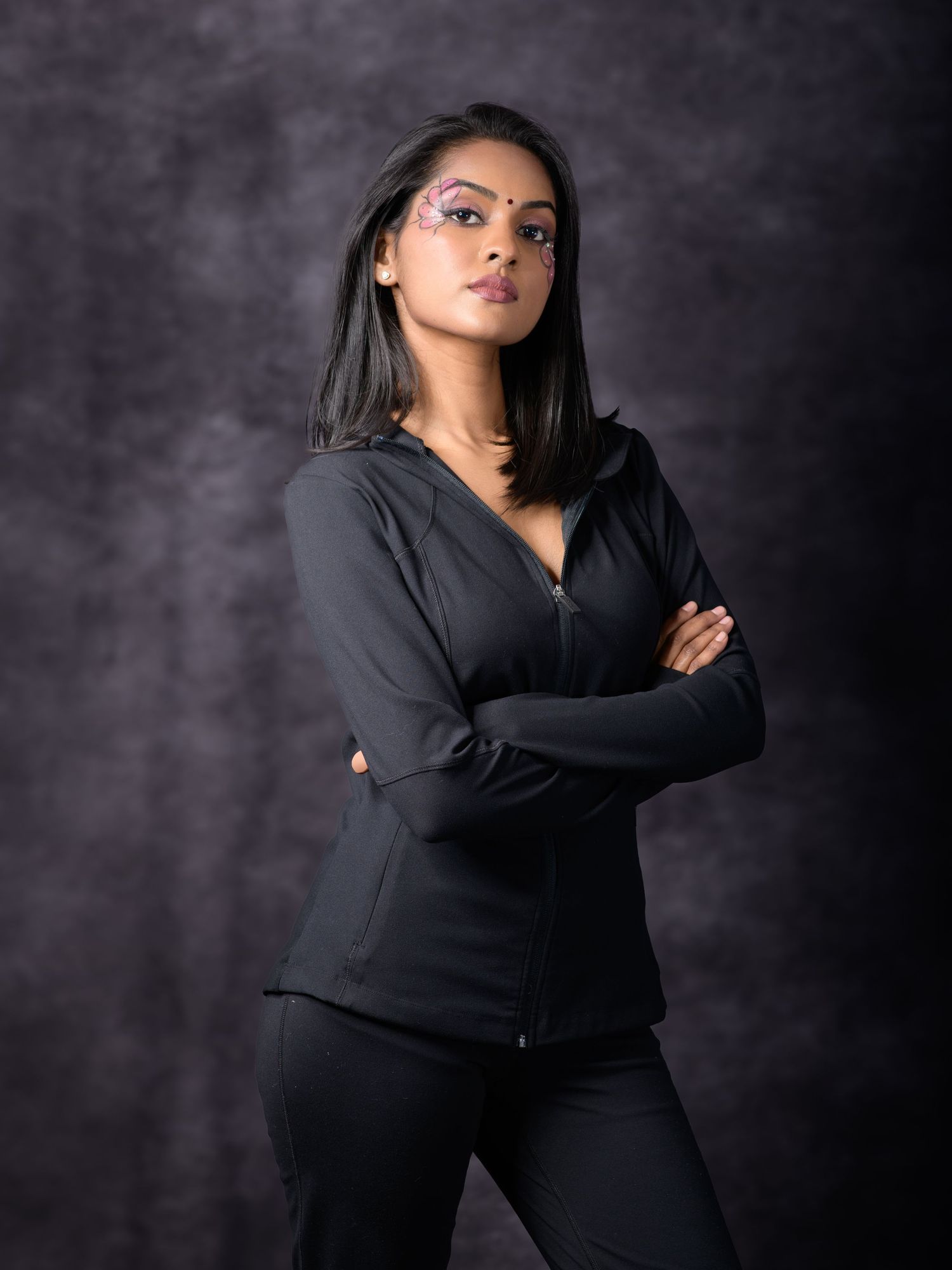Woman in black zip-up jacket with arms crossed, wearing artistic face paint, posing confidently against dark backdrop.