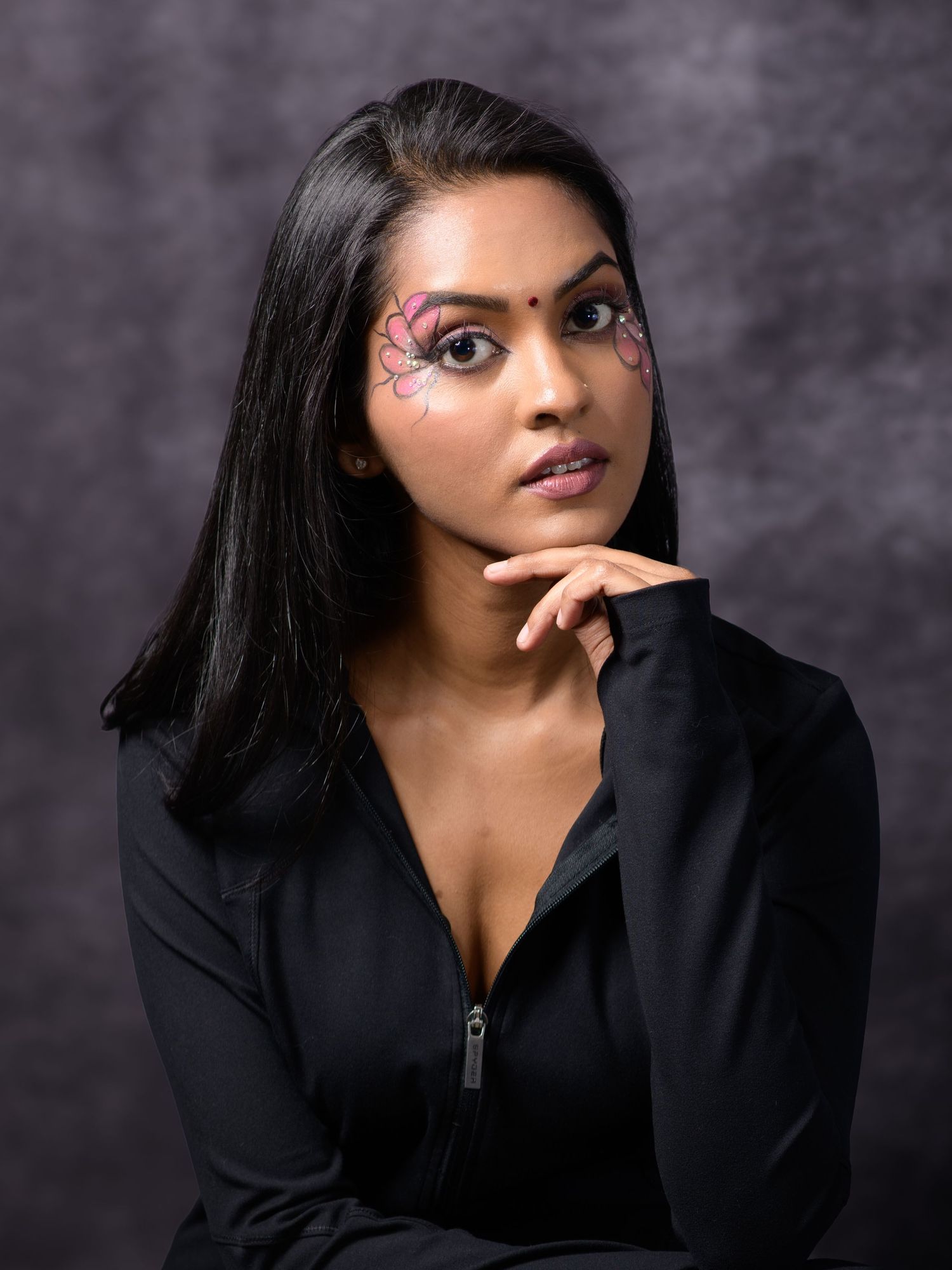 Woman with artistic butterfly face paint wearing black top poses thoughtfully against dark studio backdrop.