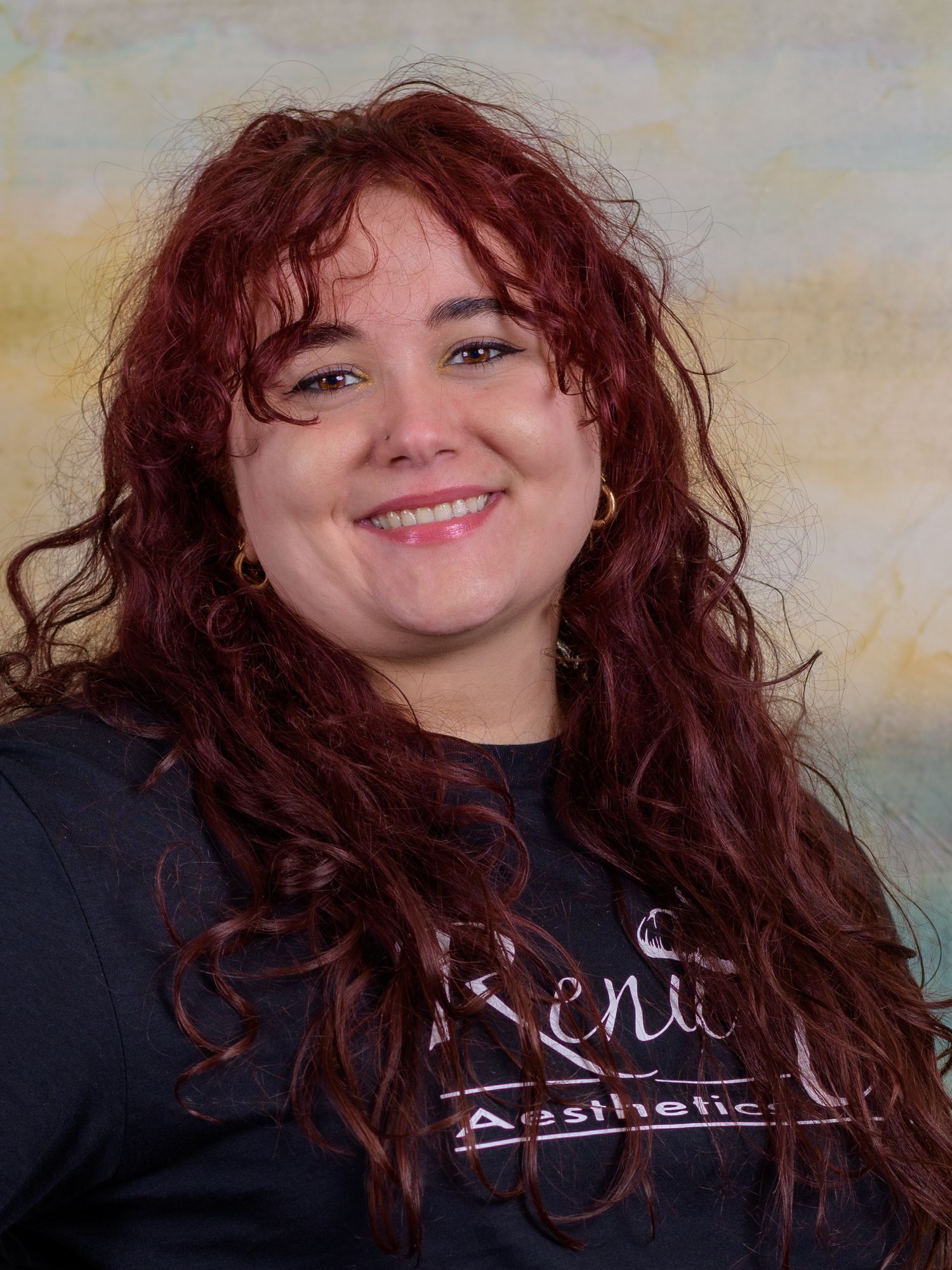 Woman with curly red hair smiling, wearing a dark Renu U Aesthetics shirt, professional headshot photo.