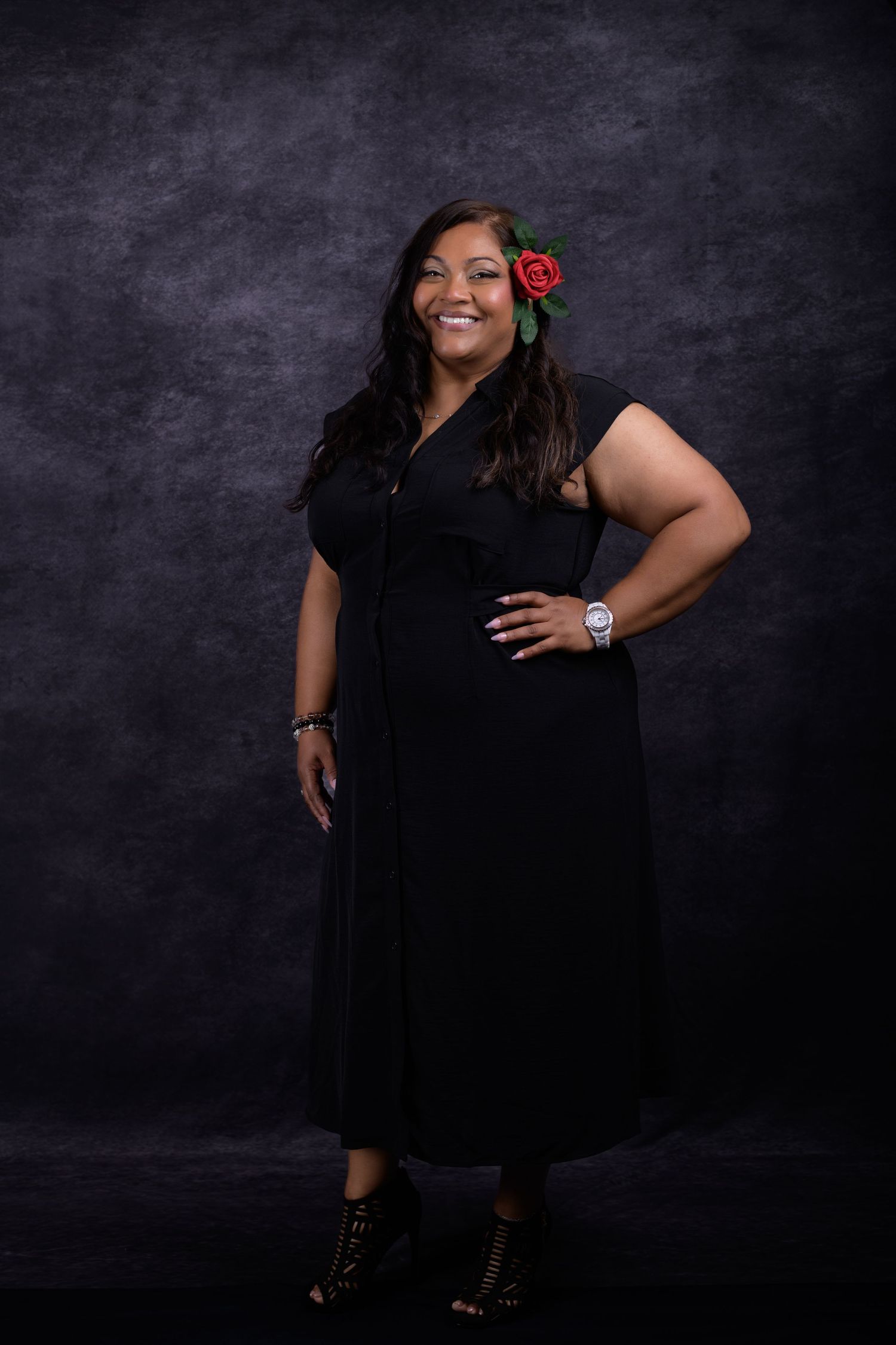 Woman in black midi dress with red rose hair accessory poses confidently against dark studio backdrop.