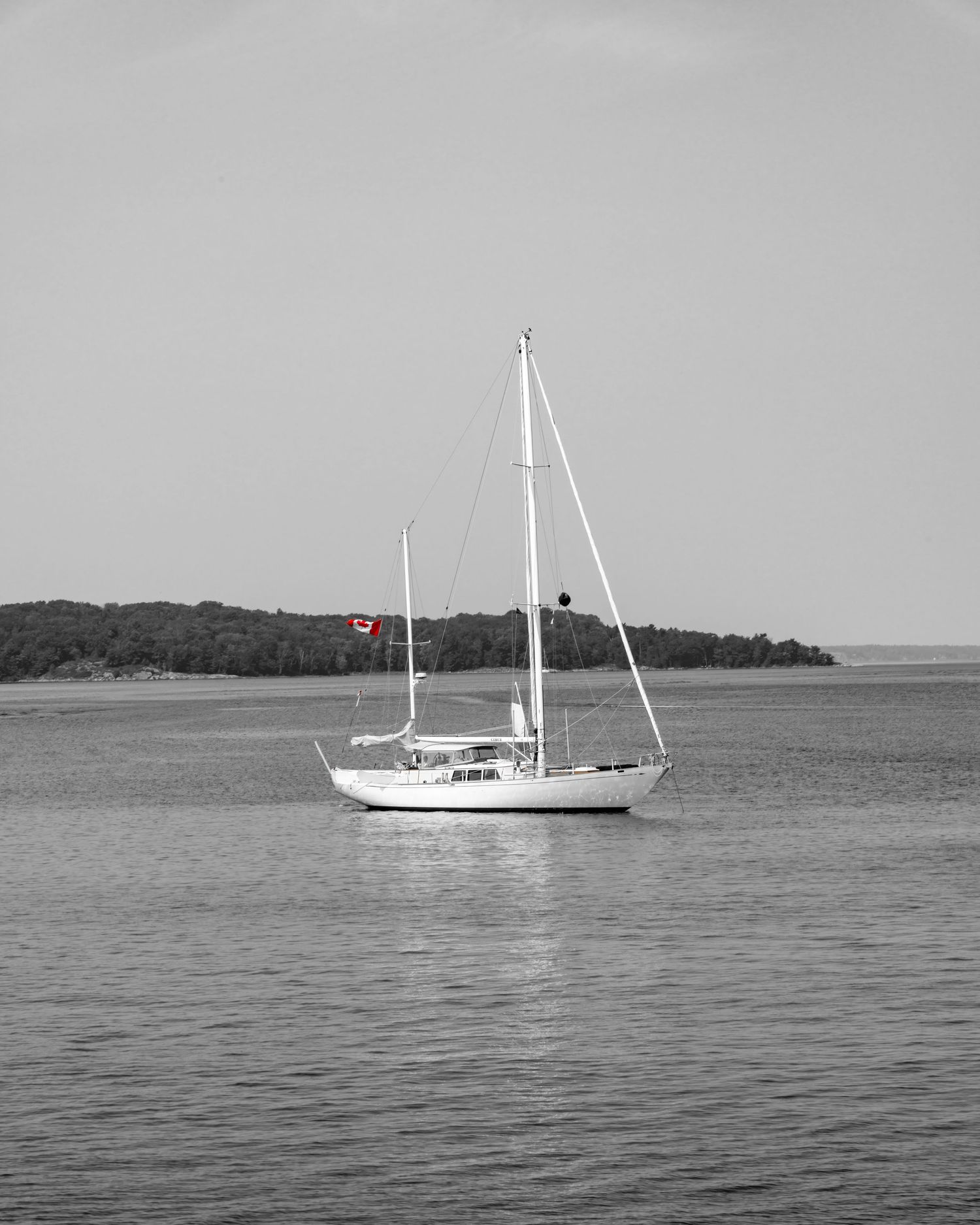 Black and white photo of a white sailboat with a red Canadian flag anchored on calm waters near a tree-lined shore.