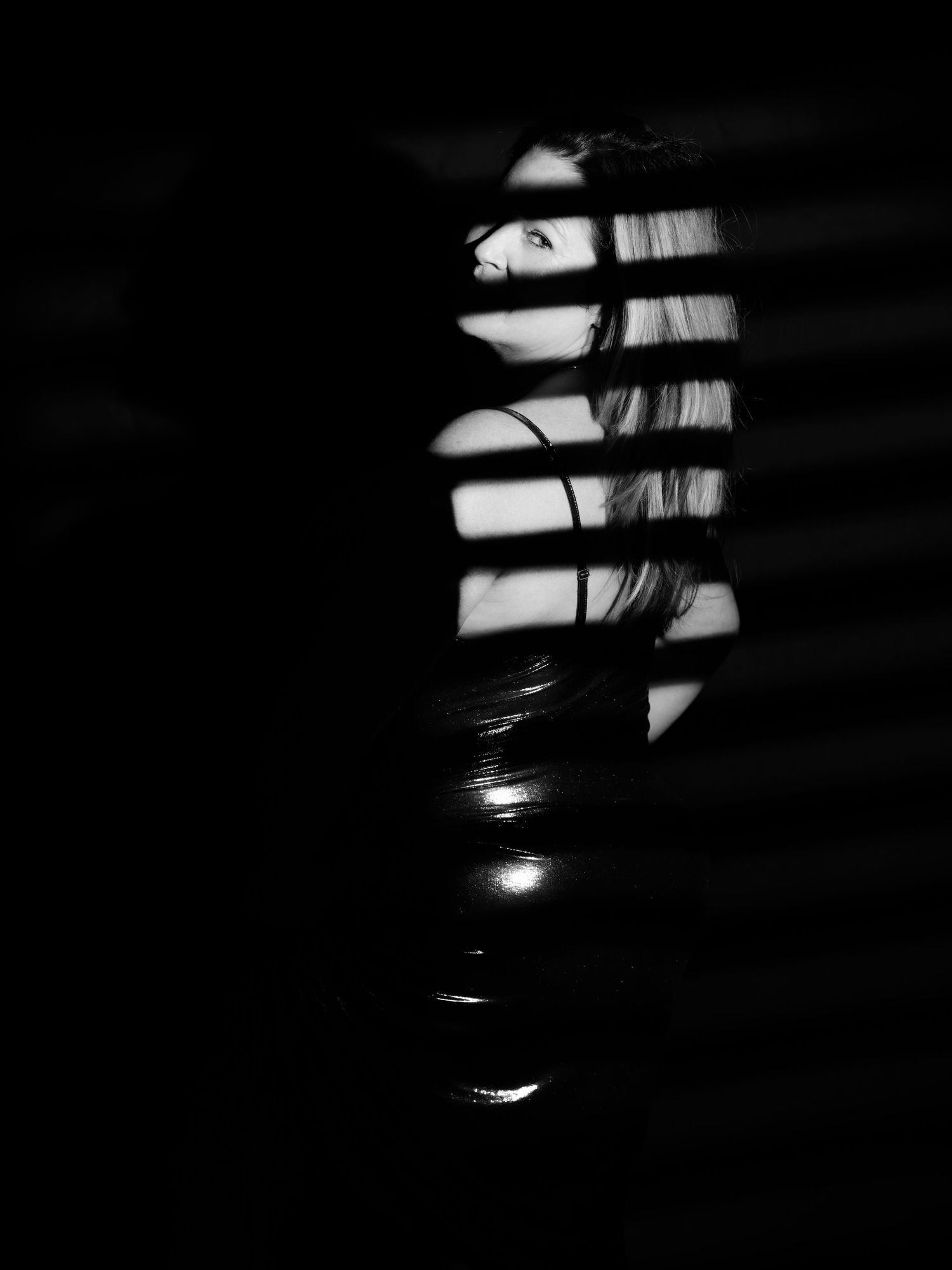 Black and white portrait of a woman illuminated by dramatic venetian blind shadow stripes in dark moody lighting.