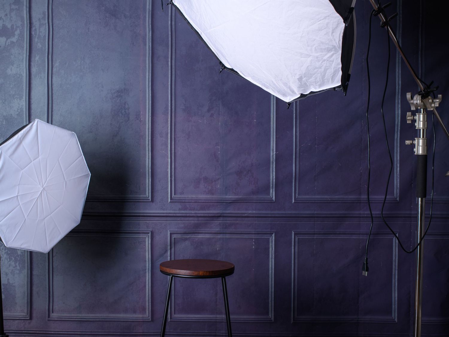 Photography studio setup with octagonal softboxes illuminating a wooden stool against a dark blue paneled wall.