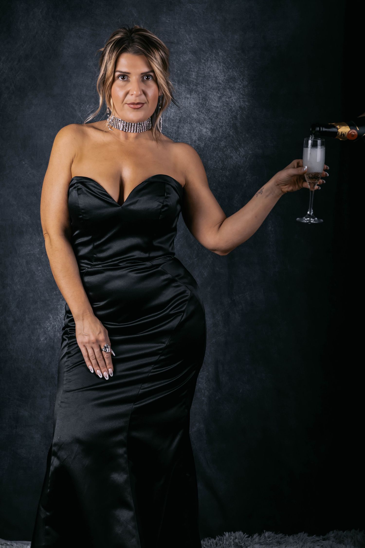 Elegant figure in black satin evening gown holds champagne glass against dark dramatic backdrop.