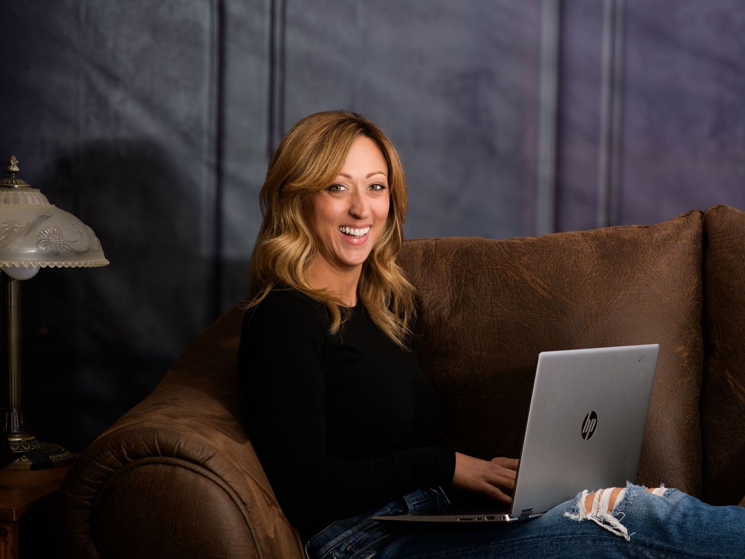 A smiling blonde woman sits on a brown leather couch using an HP laptop in a cozy, dimly lit room.