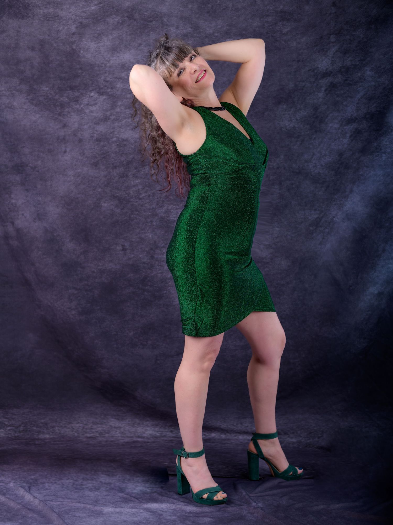 Woman posing in a green sparkly mini dress with matching heeled sandals against a gray studio backdrop.