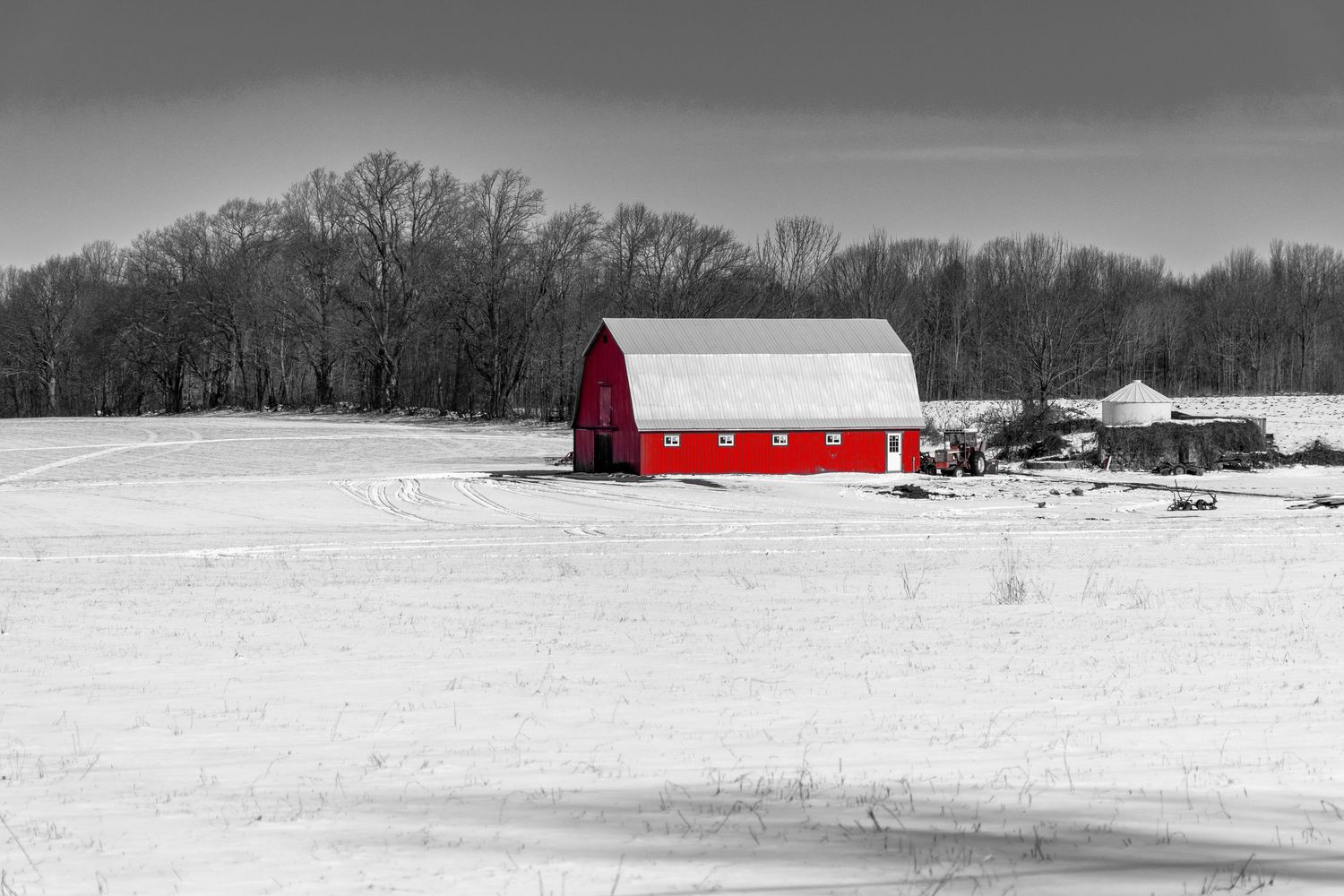 Red barn stands out in snowy winter landscape with bare trees in black and white selective color photography.