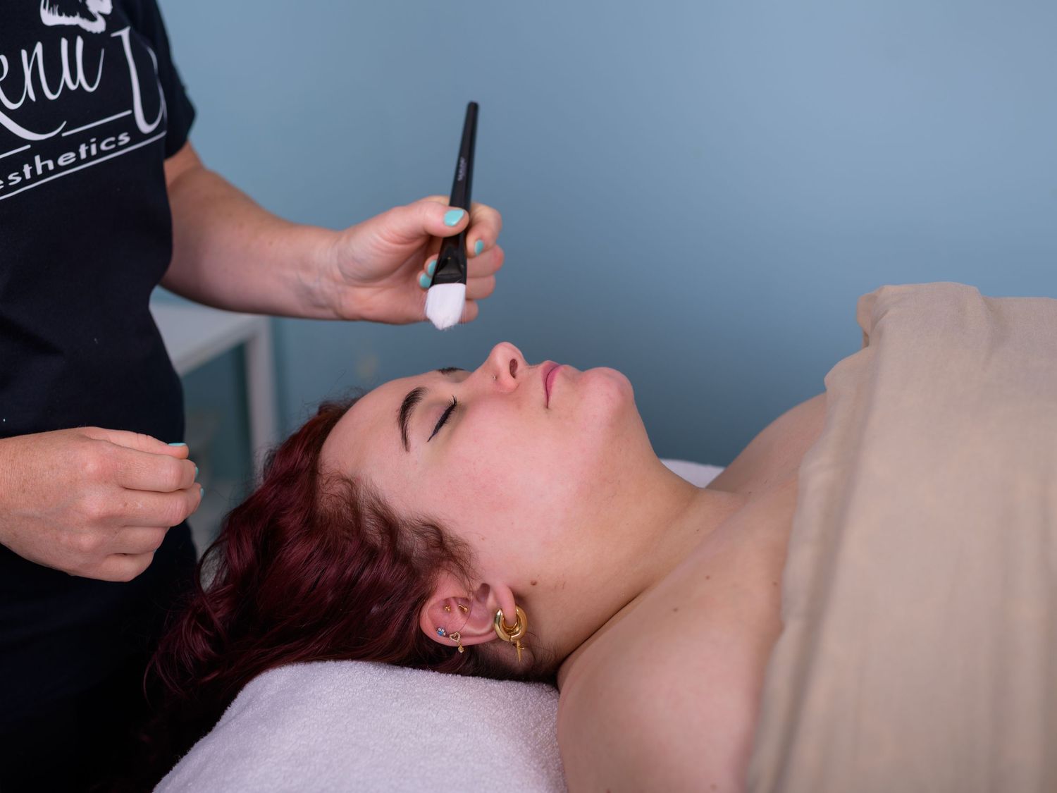 Esthetician applying facial treatment to a woman with red hair lying on a spa table.