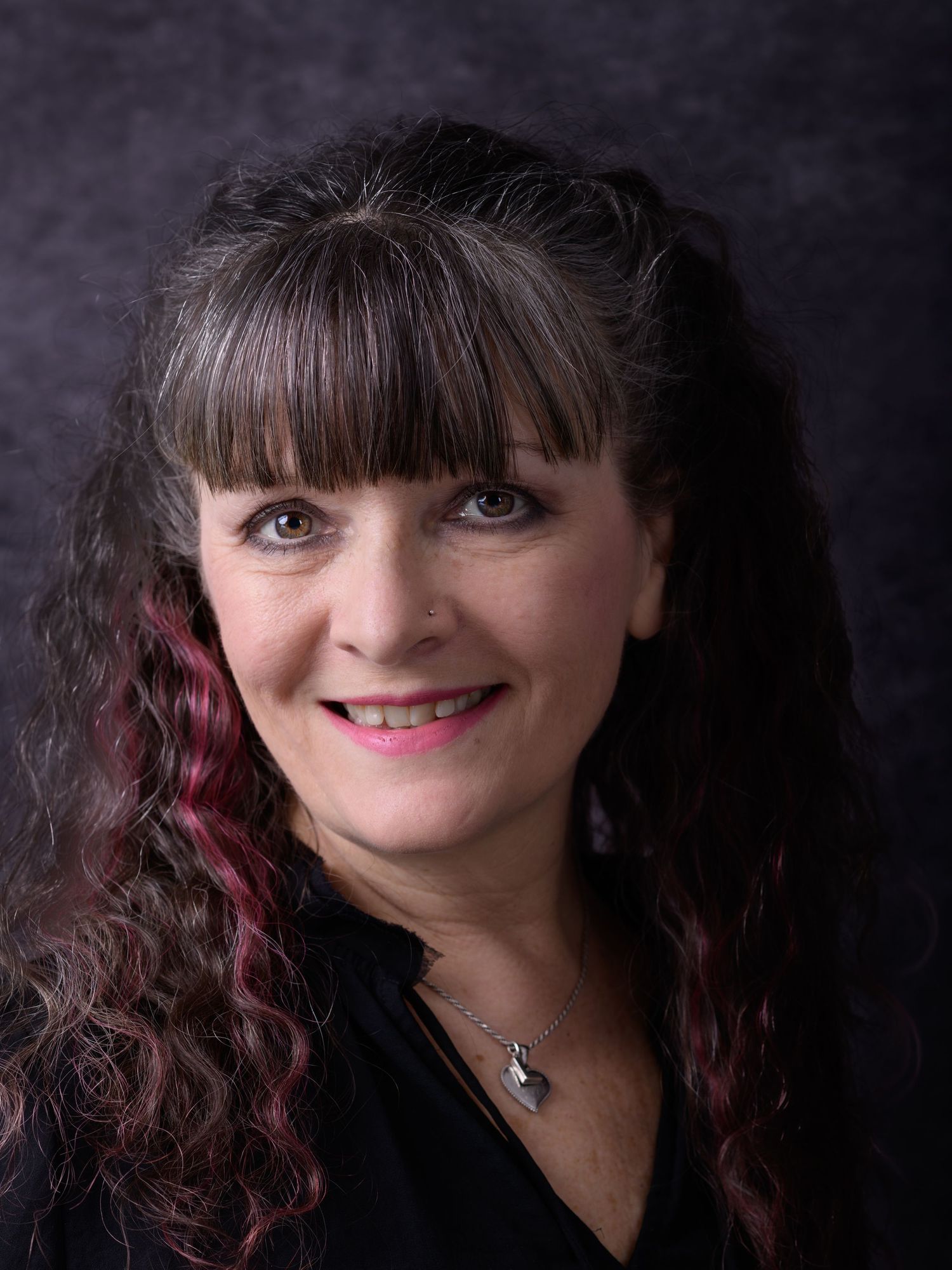 Professional headshot of a woman with dark curly hair with red highlights, bangs, and a black top against a gray background.