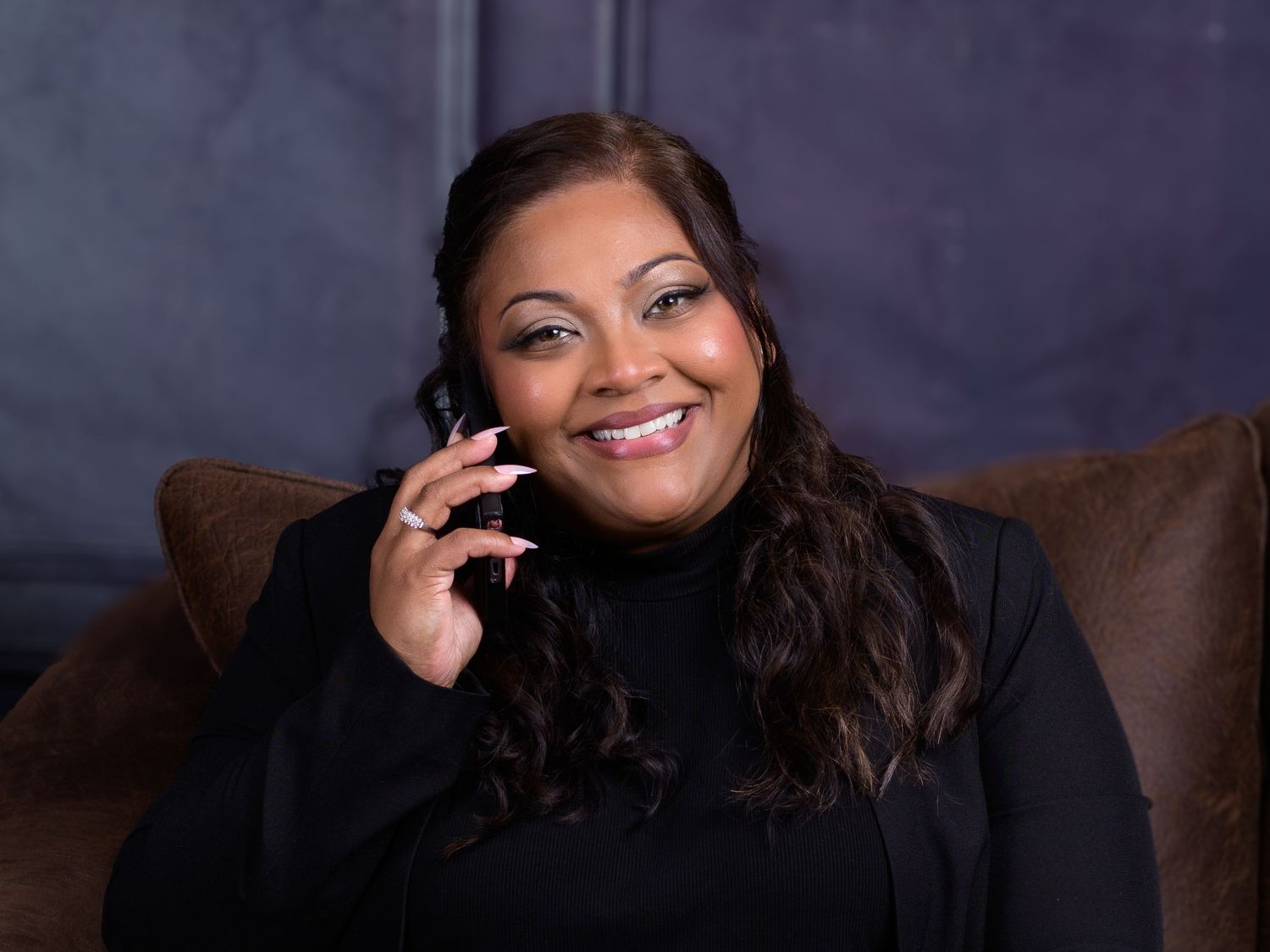 Smiling woman in black turtleneck seated on brown leather chair against dark blue background.