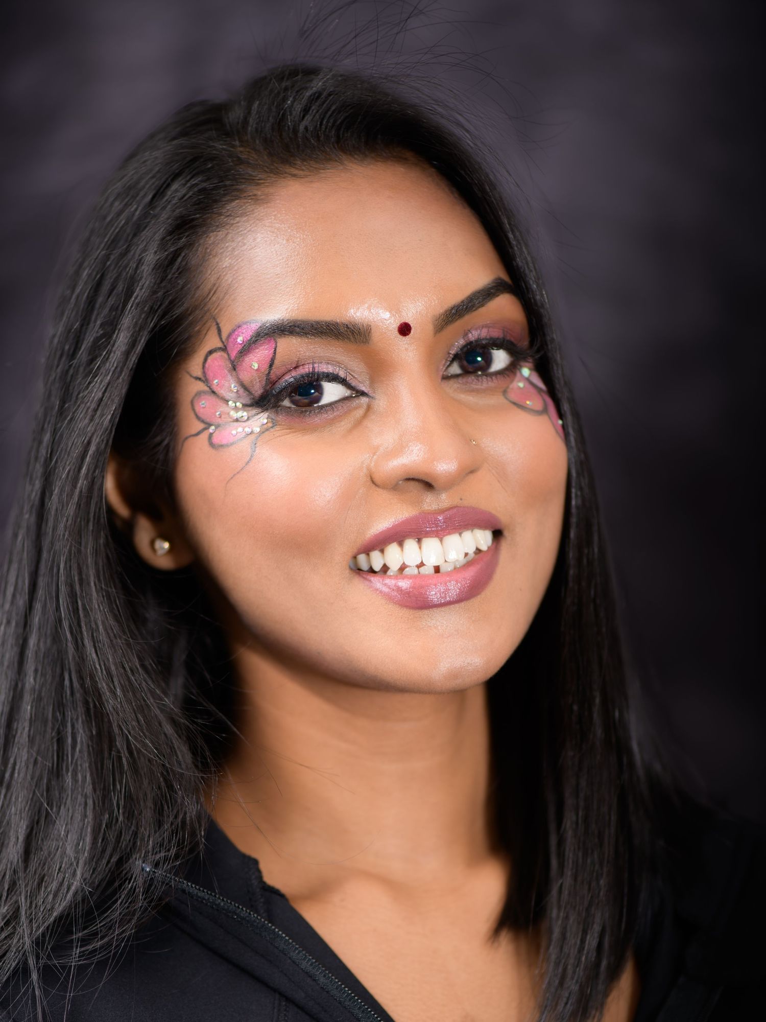 Smiling woman with pink butterfly eye makeup and bindi, wearing black top.