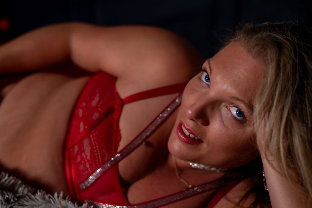 Woman in red lace lingerie posing seductively with blue eyes and blonde hair in dramatic lighting.