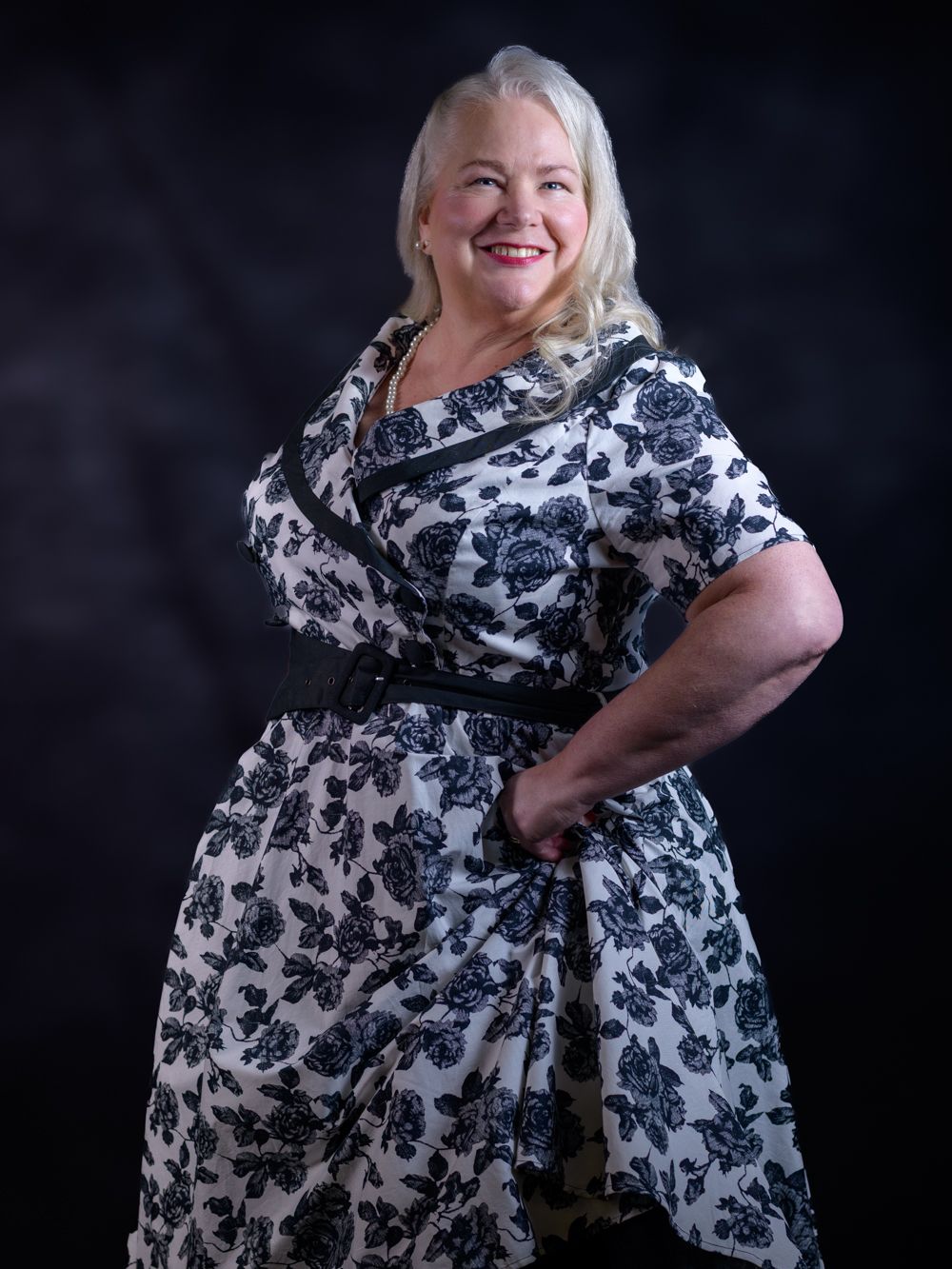 Full-length portrait of a blonde woman in a black and white floral dress posing confidently.