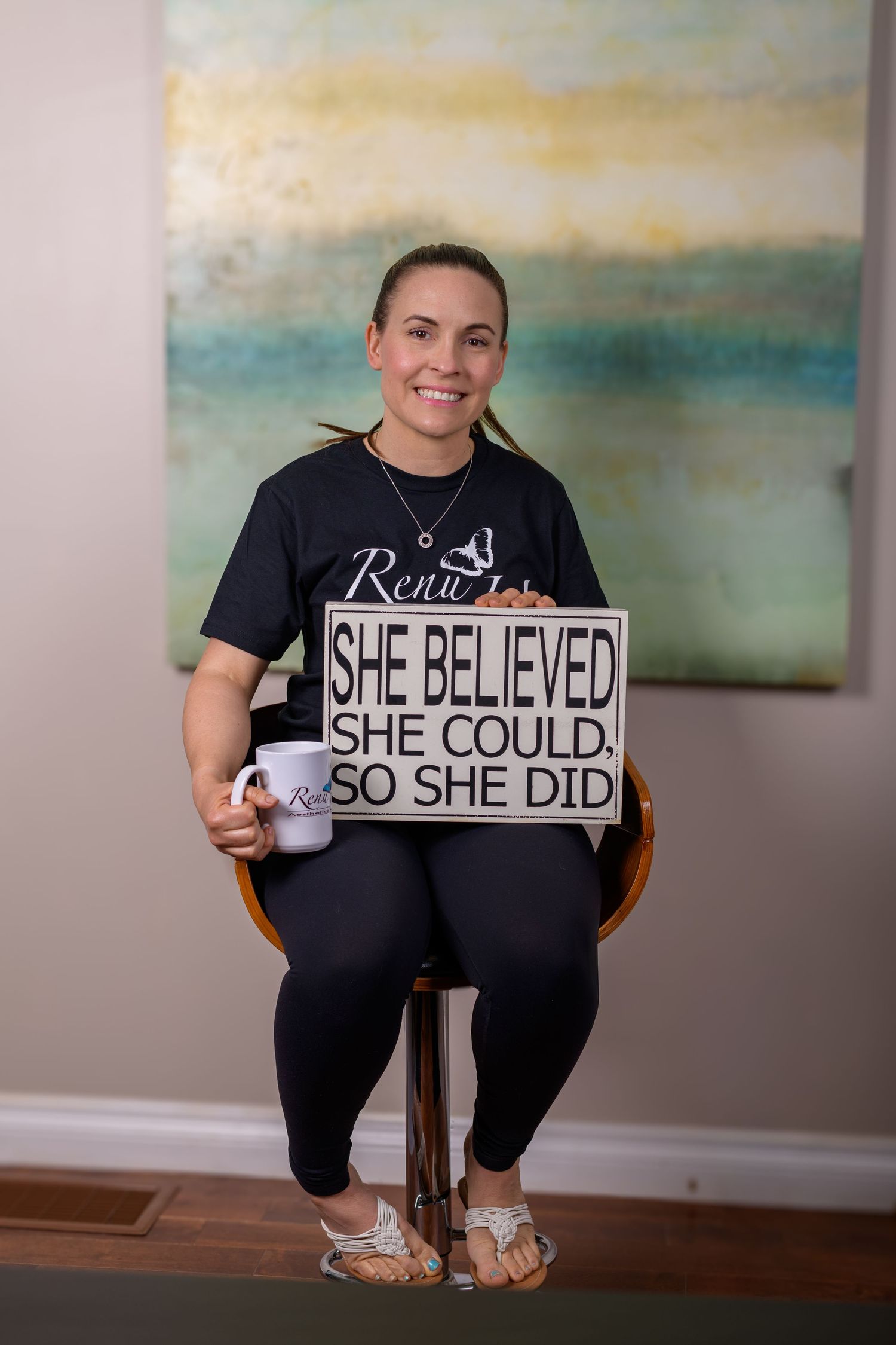 Woman in black tee holding motivational sign reading She Believed She Could So She Did, seated with coffee mug.