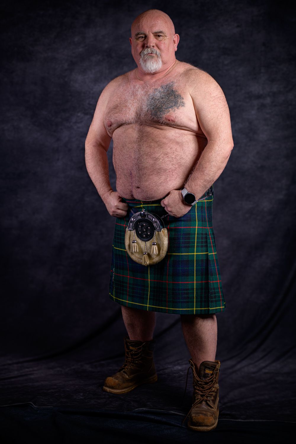 Rab Carrigan posing shirtless in a green plaid kilt and work boots against a dark textured backdrop.