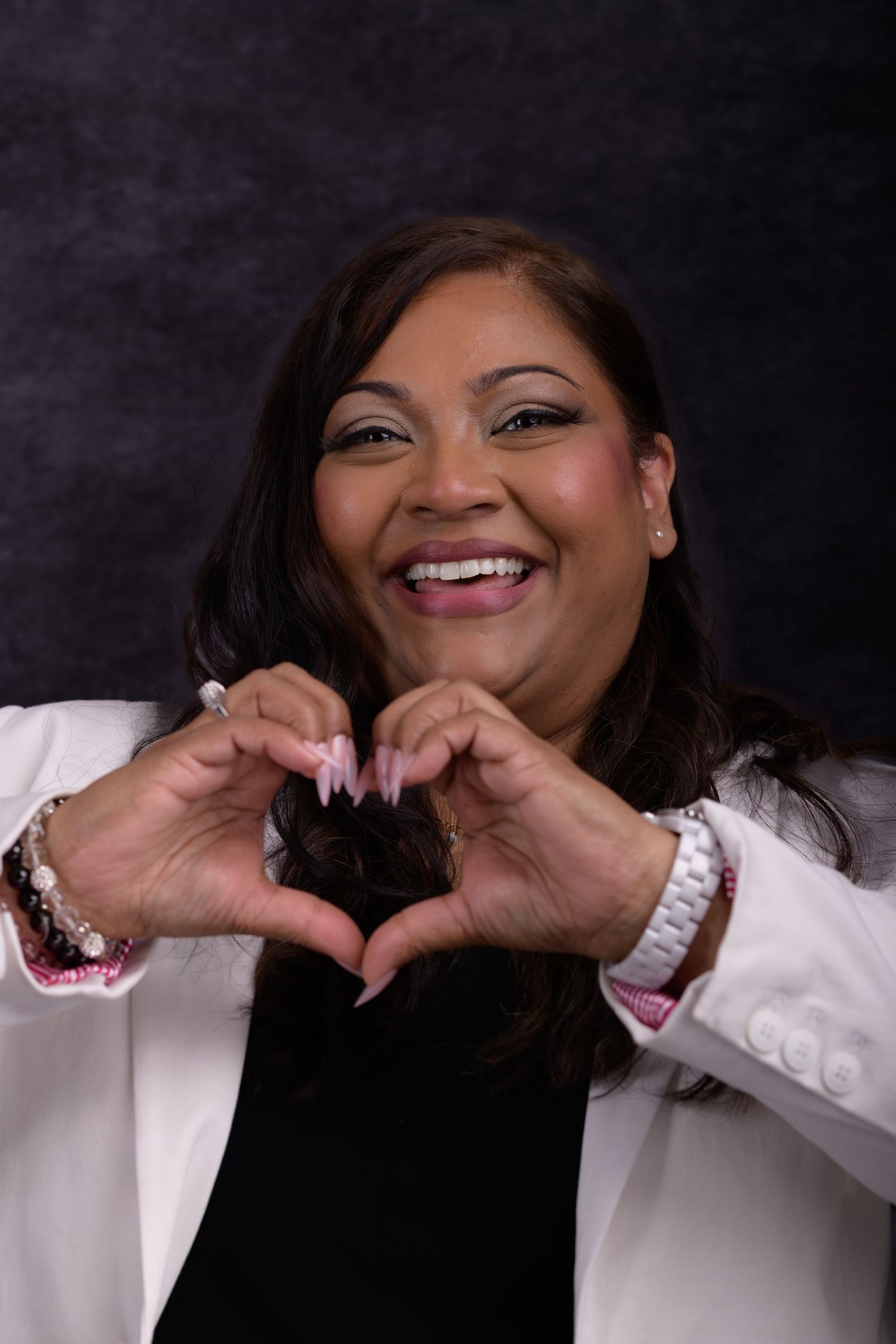 Smiling woman in white blazer making a heart shape with her hands against a dark gray studio background.