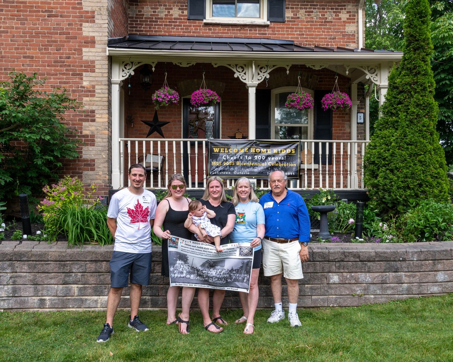 Family group photo in front of a charming brick house with hanging flower baskets and a welcome banner on a sunny day.