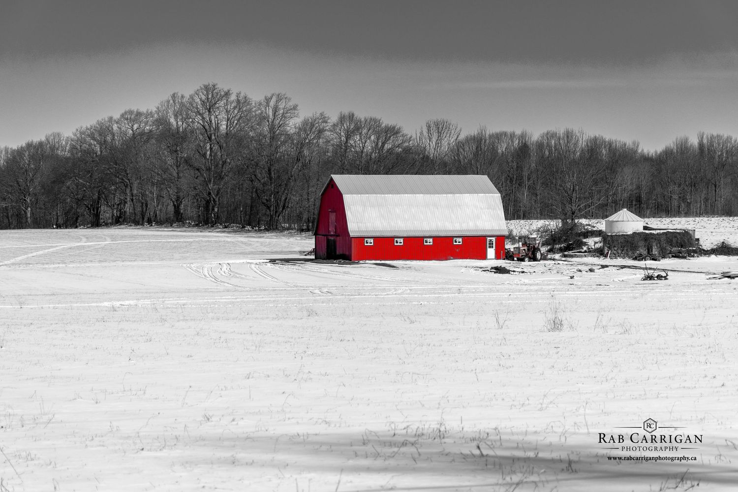 A panoramic winter landscape with a bright red barn and farm buildings surrounded by snow-covered fields and bare trees.
