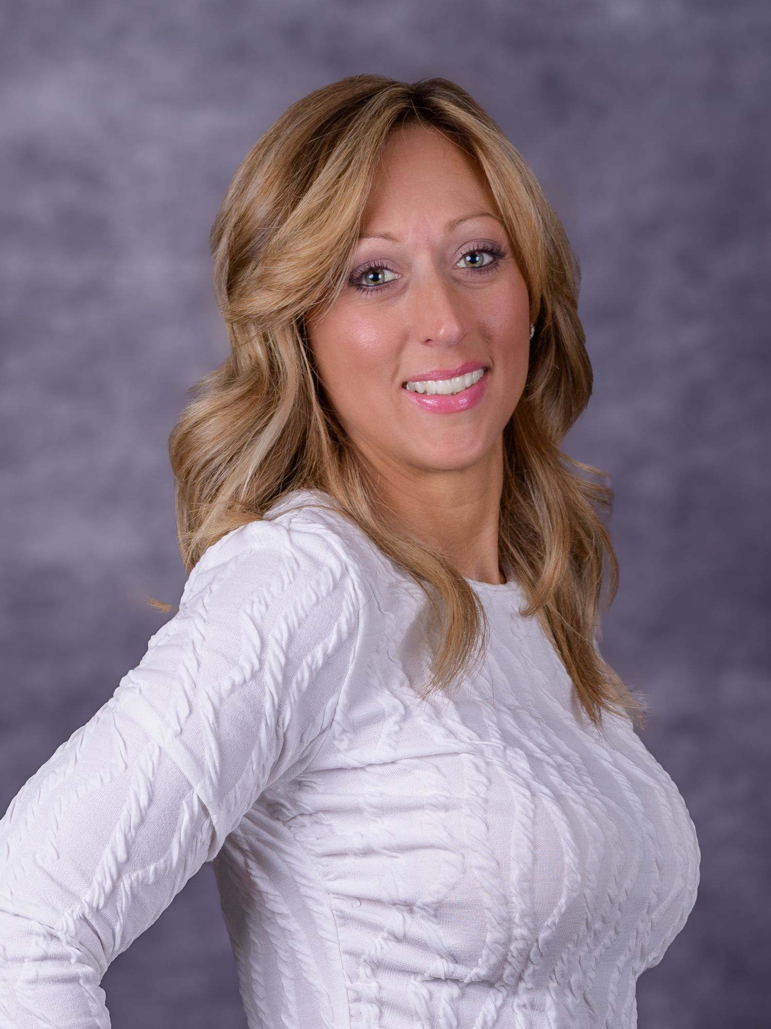 Professional headshot of a smiling blonde woman in a white textured long-sleeve top against a gray studio background.