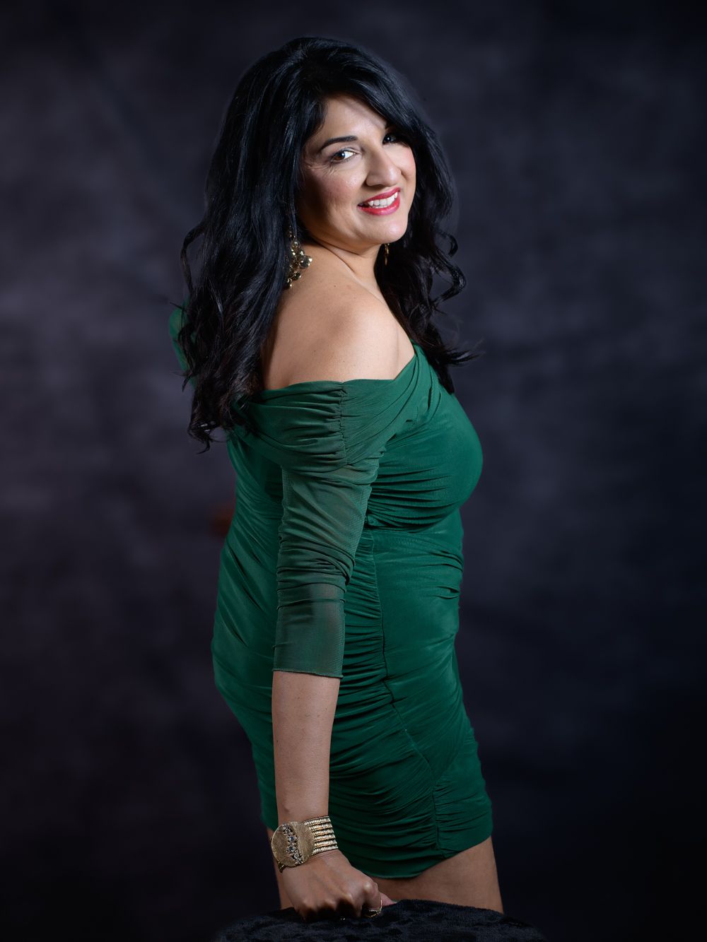 Woman in green off-shoulder ruched mini dress with black wavy hair poses confidently against dark studio background.