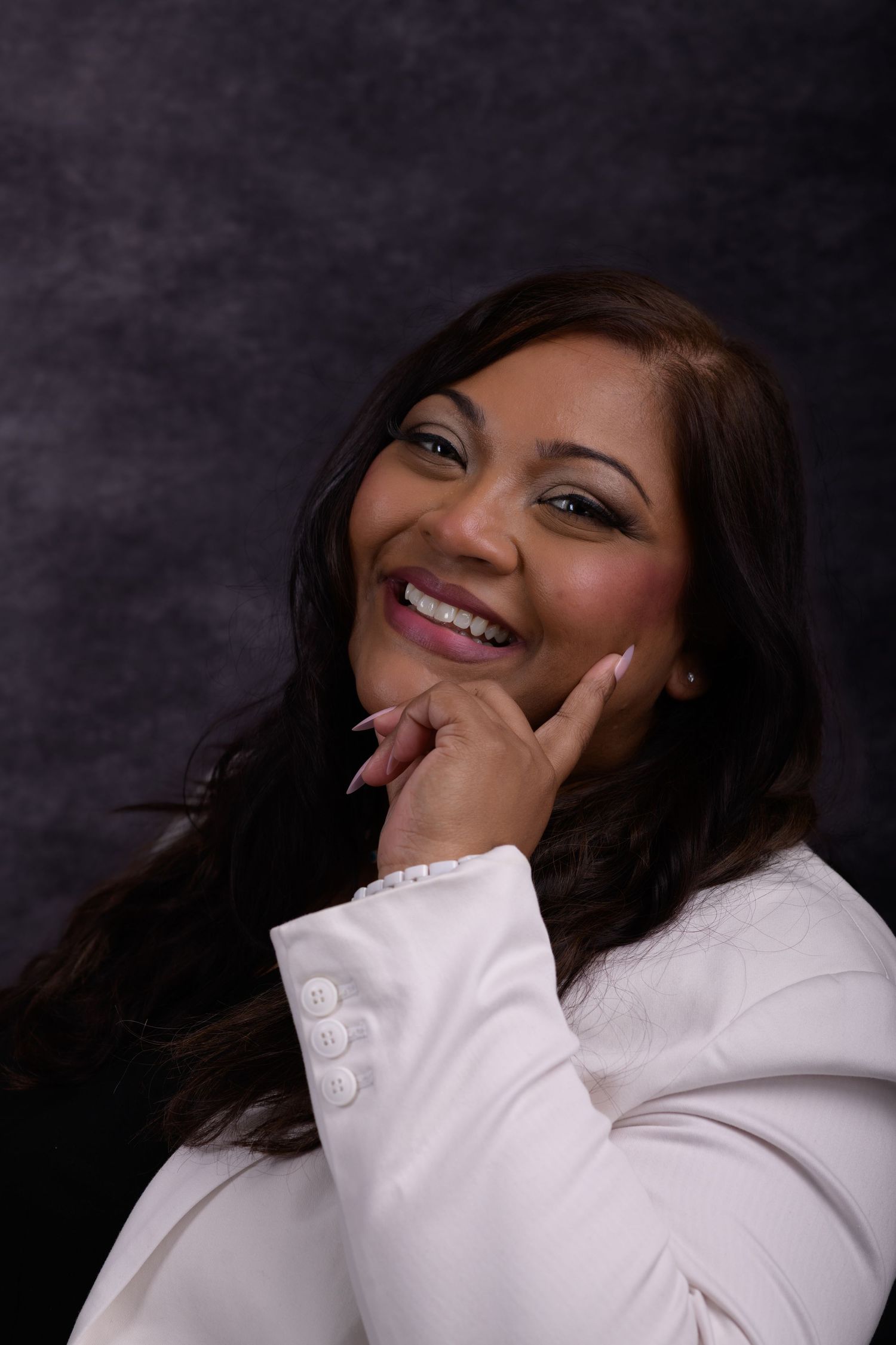 Smiling woman in white blazer posing with hand on chin against dark gray background.