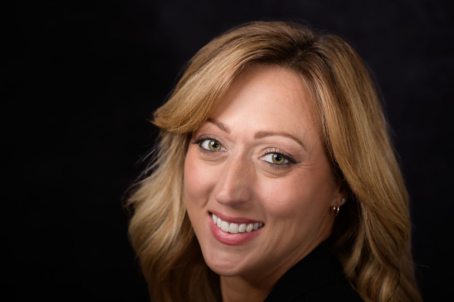 Professional headshot of a smiling blonde woman in dark attire against a dark studio background.