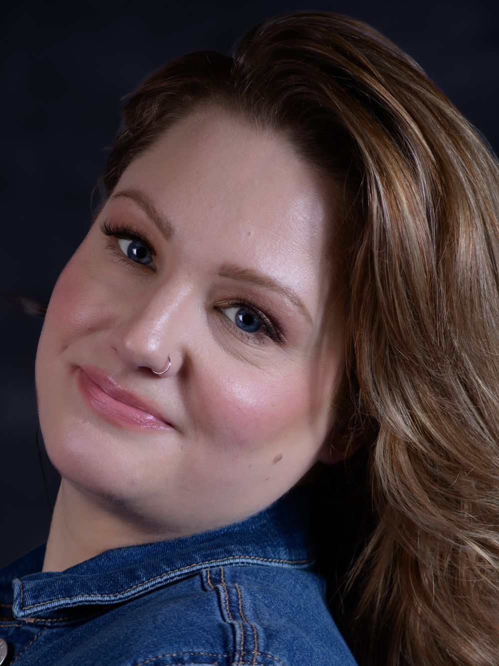 A smiling woman with auburn hair and a nose ring wearing a denim jacket against a dark background.