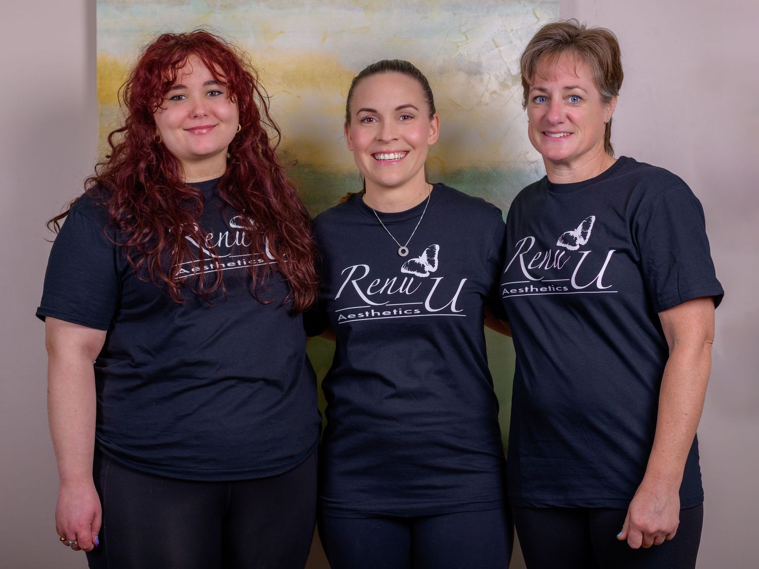 Three women wearing matching navy Renu U Aesthetics branded t-shirts smile together for a professional group photo.