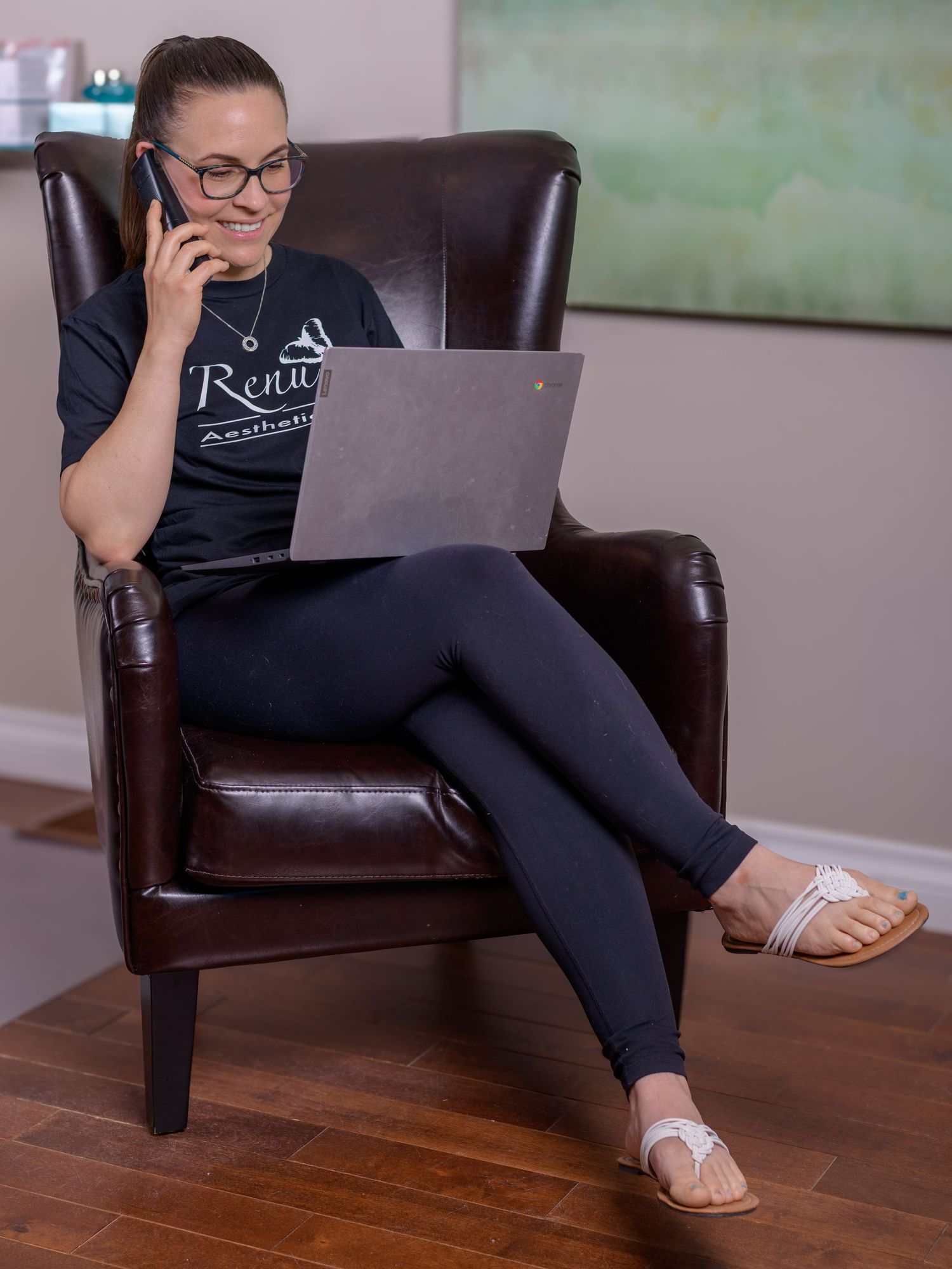 Woman in Renu U Aesthetics shirt sits in leather chair, multitasking on laptop while talking on phone.