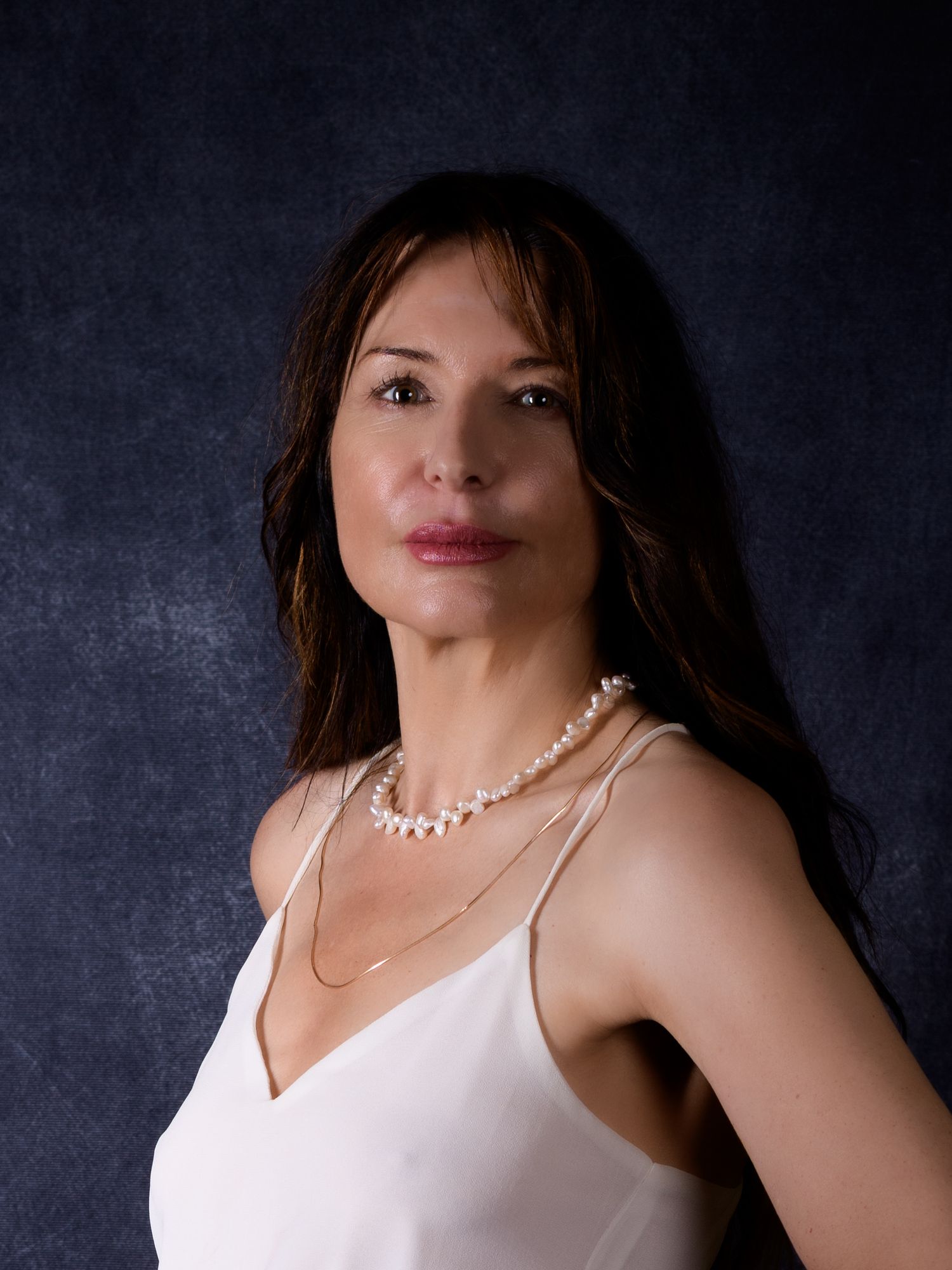 Woman with long dark hair wearing a white dress and pearl necklace poses against a dark blue background.