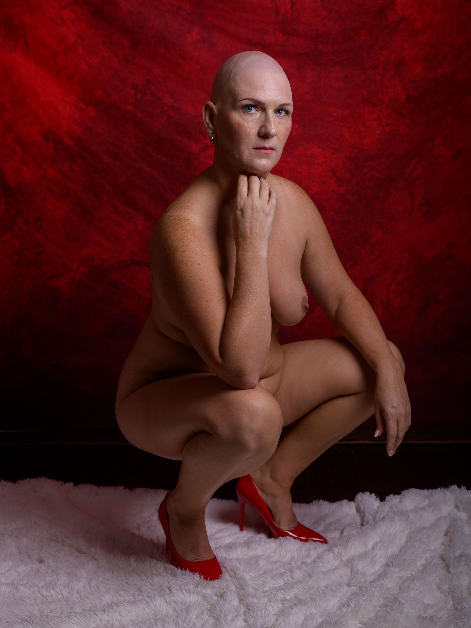 Bald woman posing nude in a squat position wearing red heels against a vibrant red backdrop on white fur.