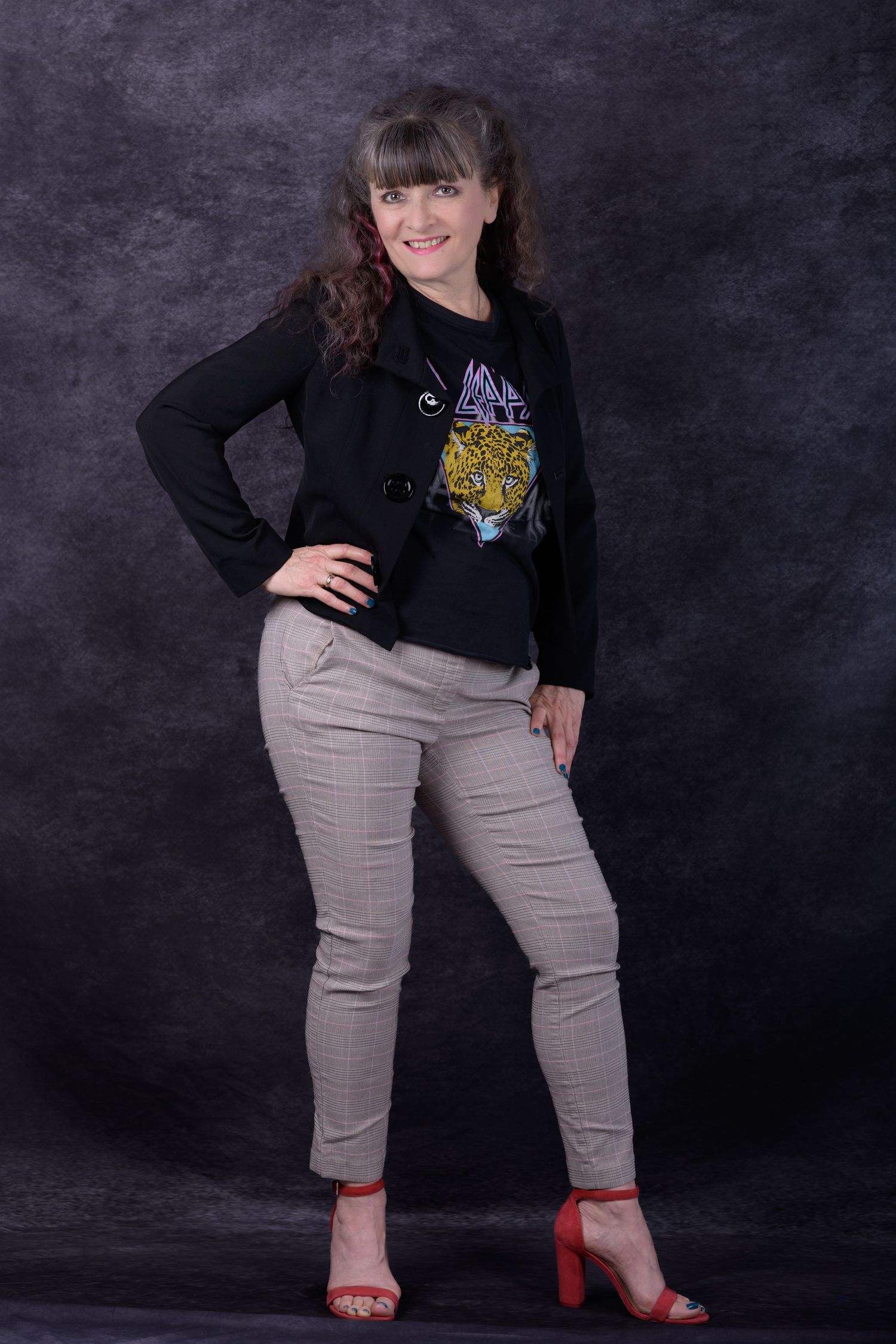 Woman posing confidently in black blazer, tiger graphic tee, gray pants, and red heeled sandals against dark studio backdrop.
