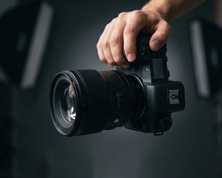 A hand holding a professional DSLR camera with a large lens against a dark studio background.