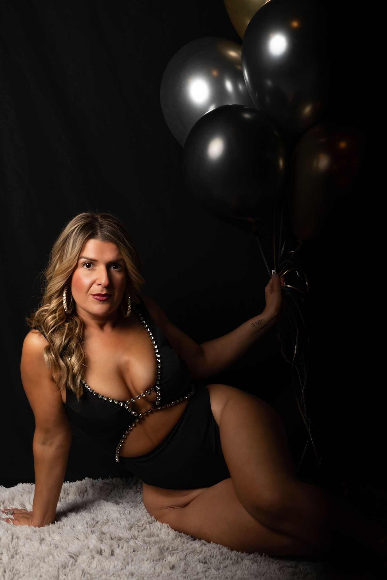 Blonde woman in black embellished swimsuit poses on white fur rug holding black and gold balloons.