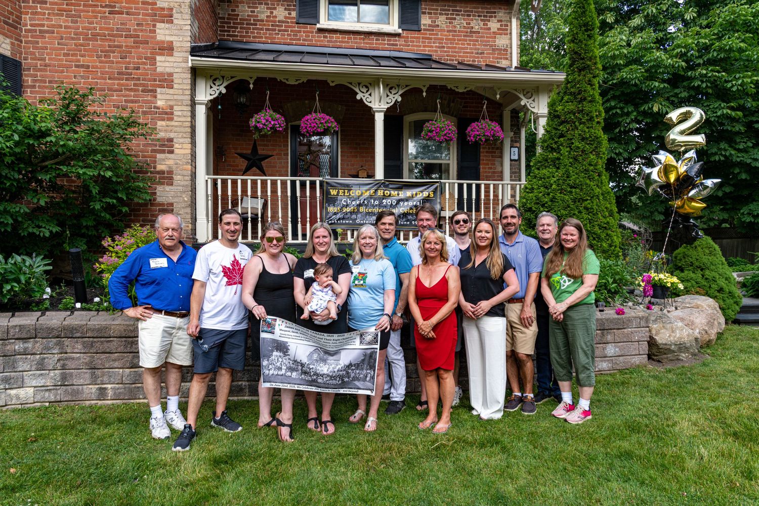 Group of people posing in front of a brick house with hanging flower baskets and a gold number 2 balloon.
