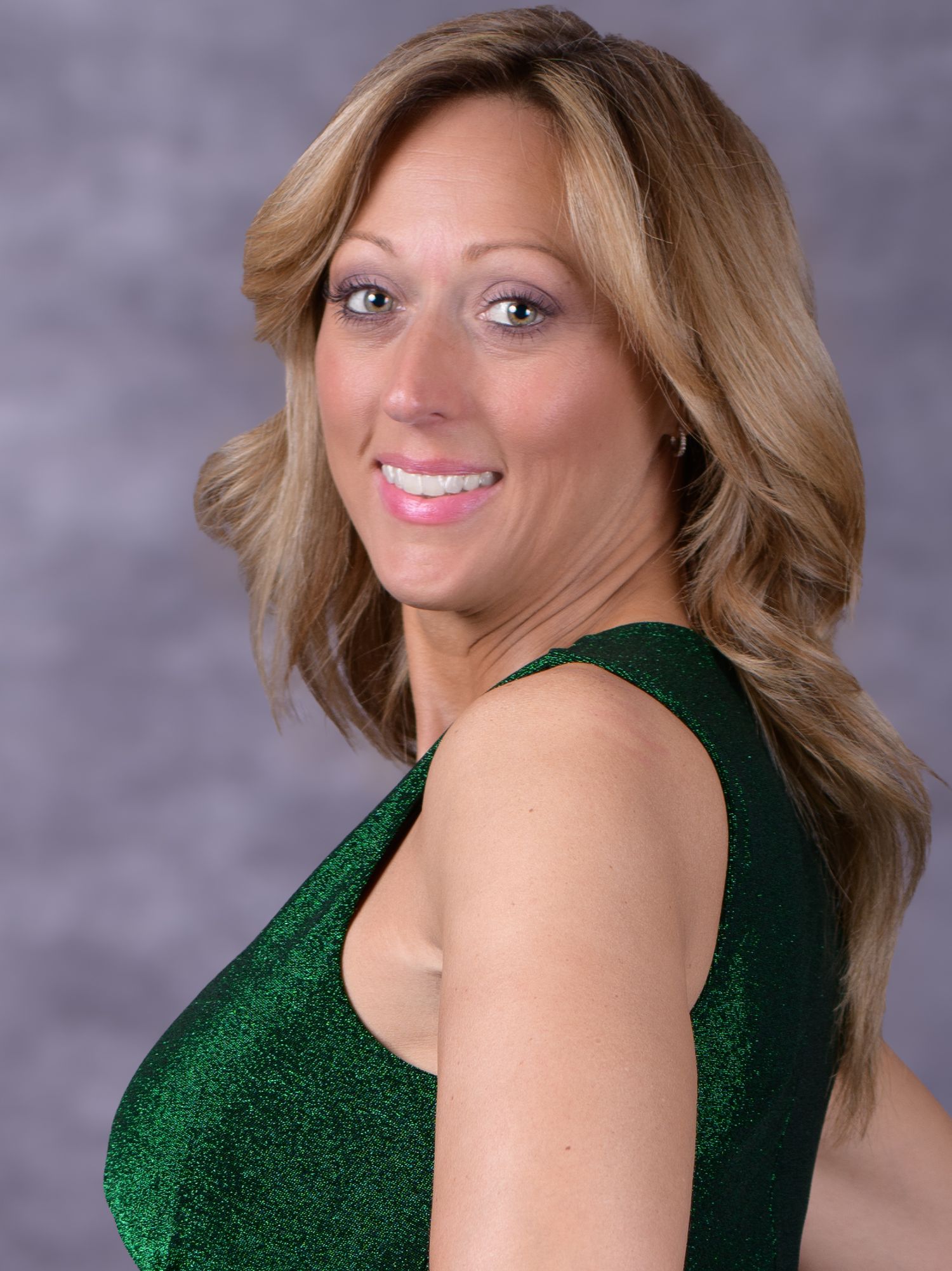 Smiling woman with blonde hair wearing a green sparkly sleeveless dress against a gray background.