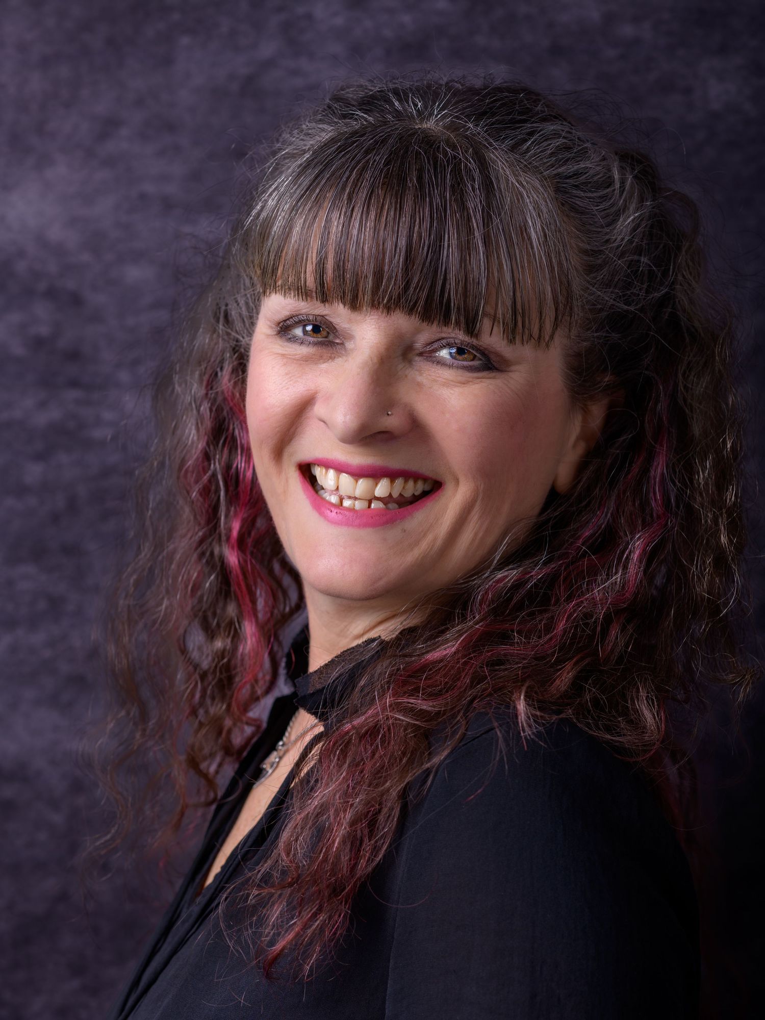Woman with dark curly hair and red highlights smiling in black outfit against gray background.