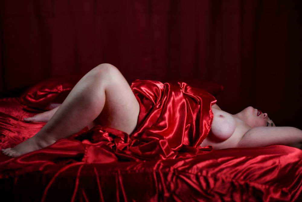 A woman draped in red satin fabric reclines on a red silk bed in a dramatic, artistic boudoir photograph.