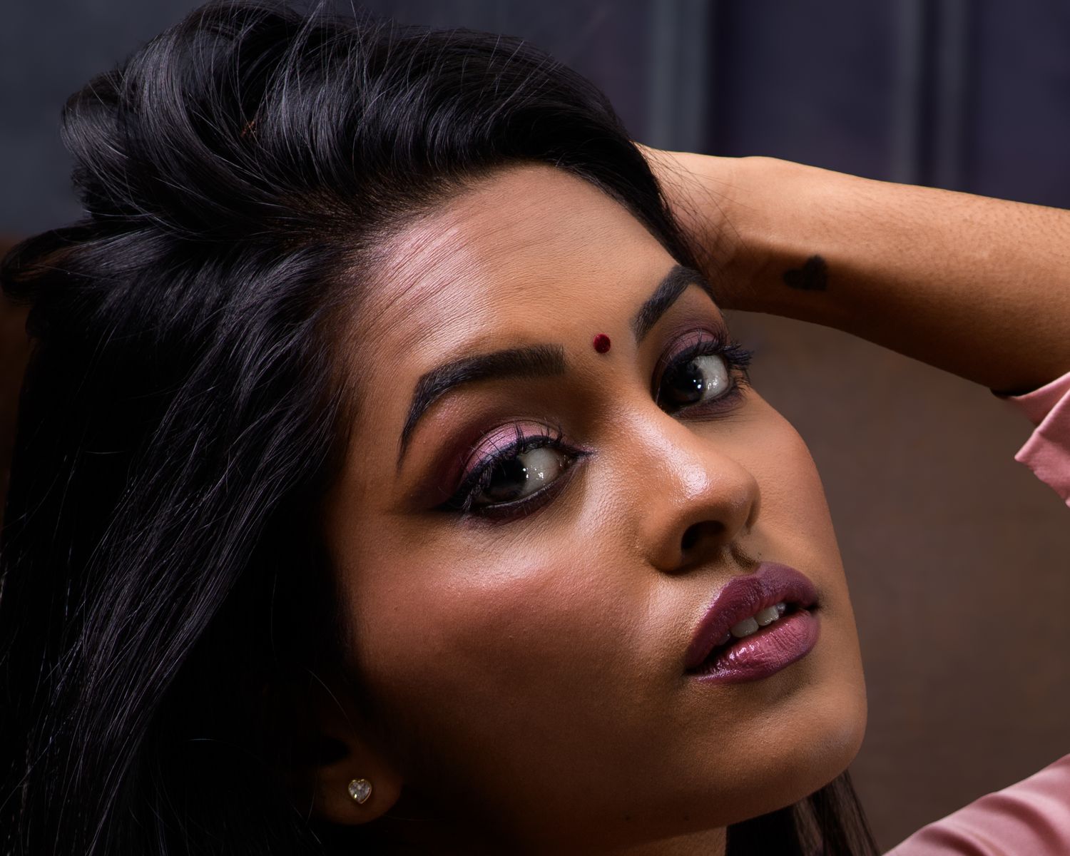 A young South Asian woman with bold eye makeup, bindi, and dark hair poses gracefully with her hand raised near her head.