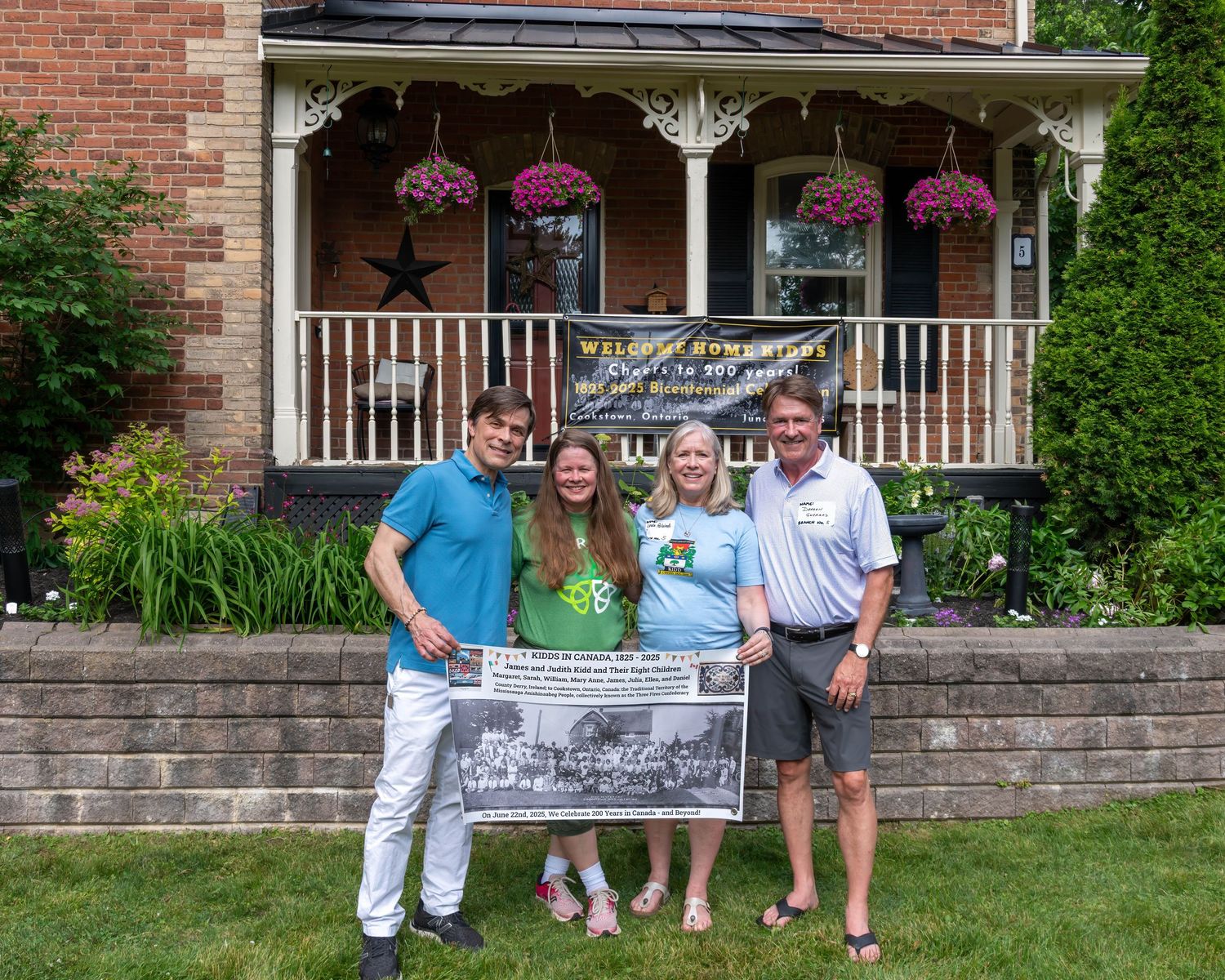 Four people holding a historical banner stand in front of a charming brick home with a decorated porch and hanging flower baskets.