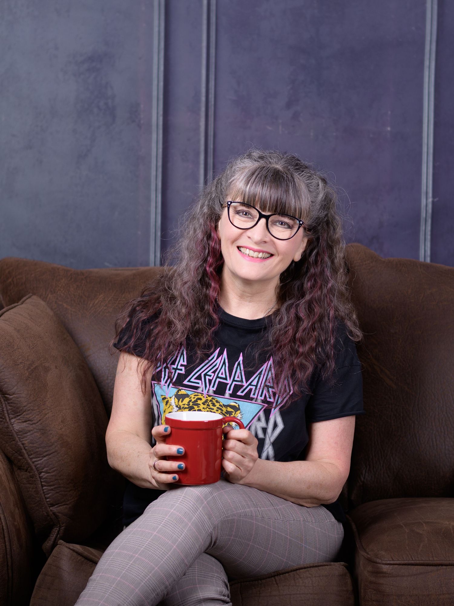 Woman with curly gray hair and glasses smiles on brown couch, wearing Def Leppard tee, holding red popcorn bucket.