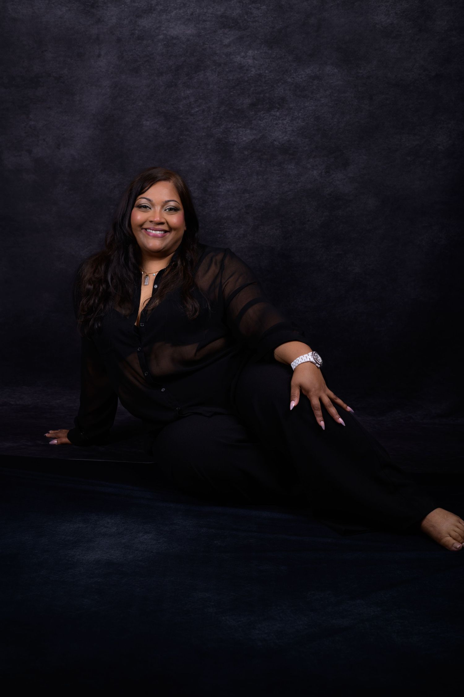 A smiling woman in black sheer top and pants poses seated on a dark studio floor against a moody gray backdrop.