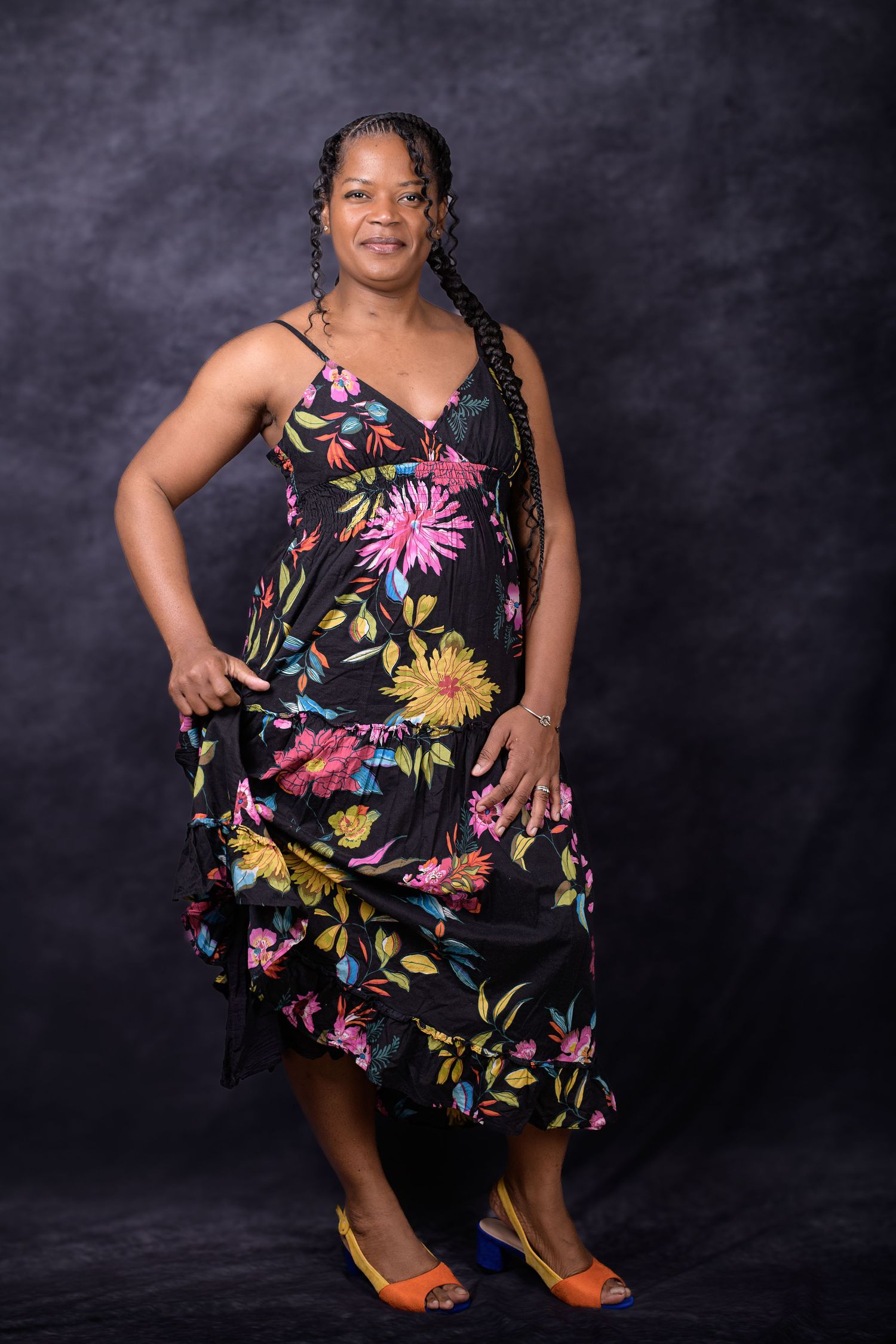 A woman in a black floral maxi dress with colorful flowers poses confidently in mismatched shoes against a dark backdrop.