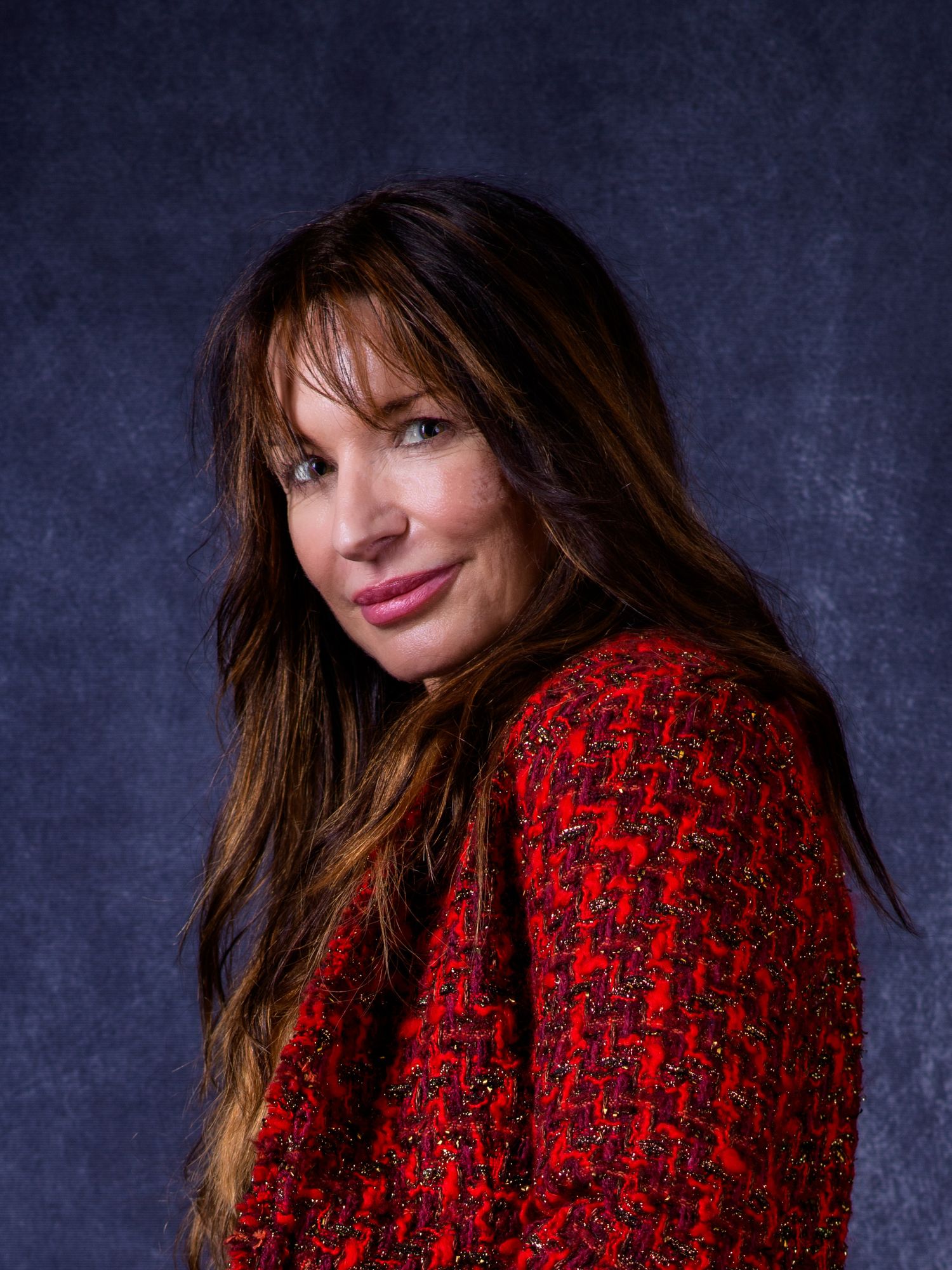 Woman with long brunette hair wearing a red tweed jacket, posing against a dark blue studio background.