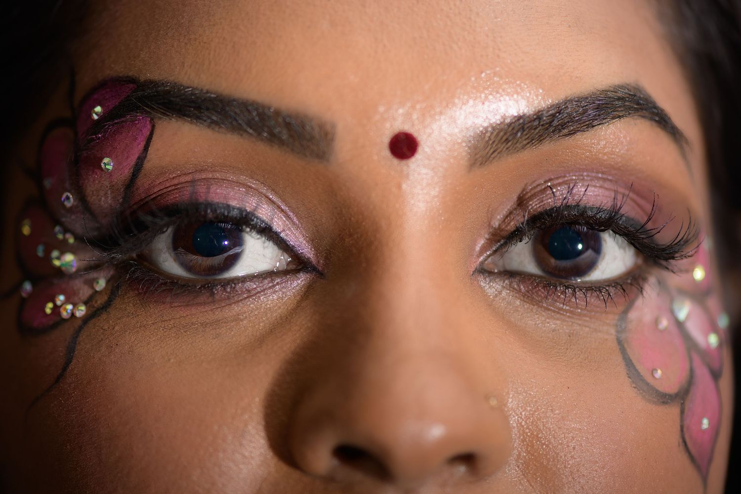 Close-up of a woman with vibrant butterfly face paint, glitter eye makeup, red bindi, and dramatic lashes.