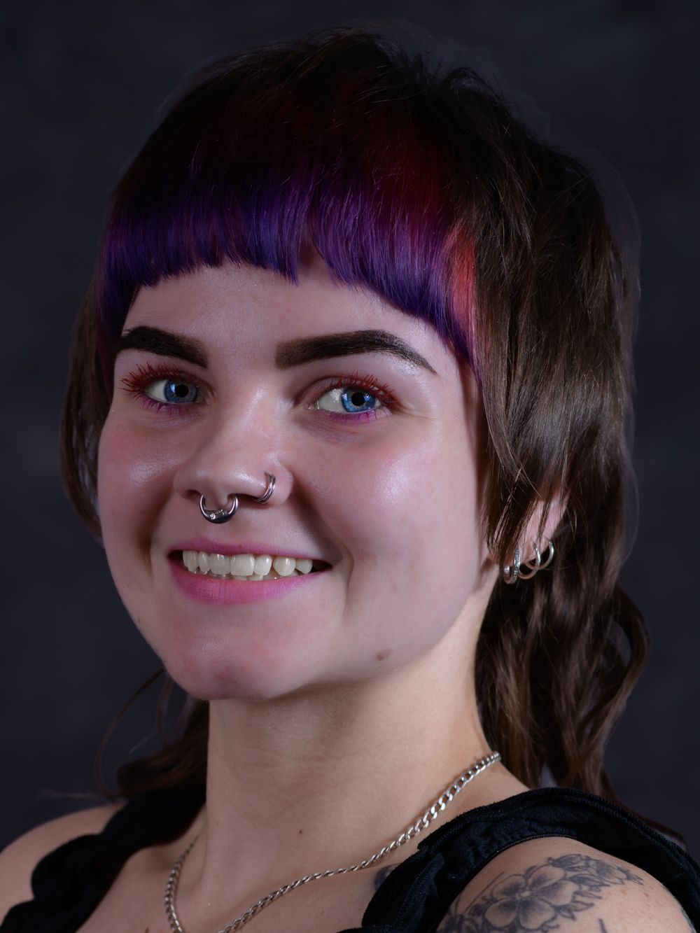 Close-up portrait of a young woman with purple and red bangs, nose ring, and tattoos smiling.