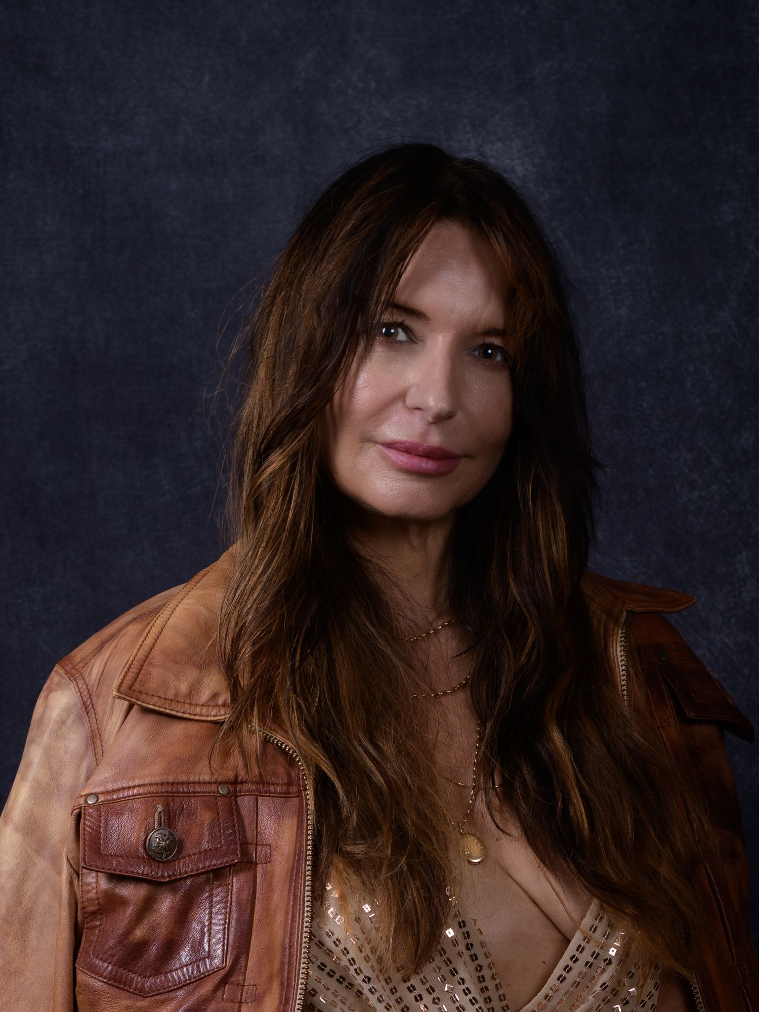 Woman with long brown hair wearing a tan leather jacket and layered necklaces poses against a dark background.