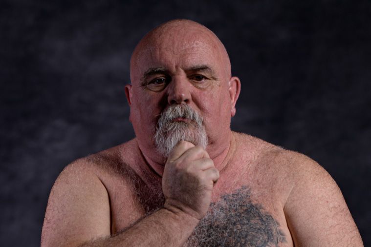 A shirtless person with a gray beard poses thoughtfully with hand on chin against a dark background.