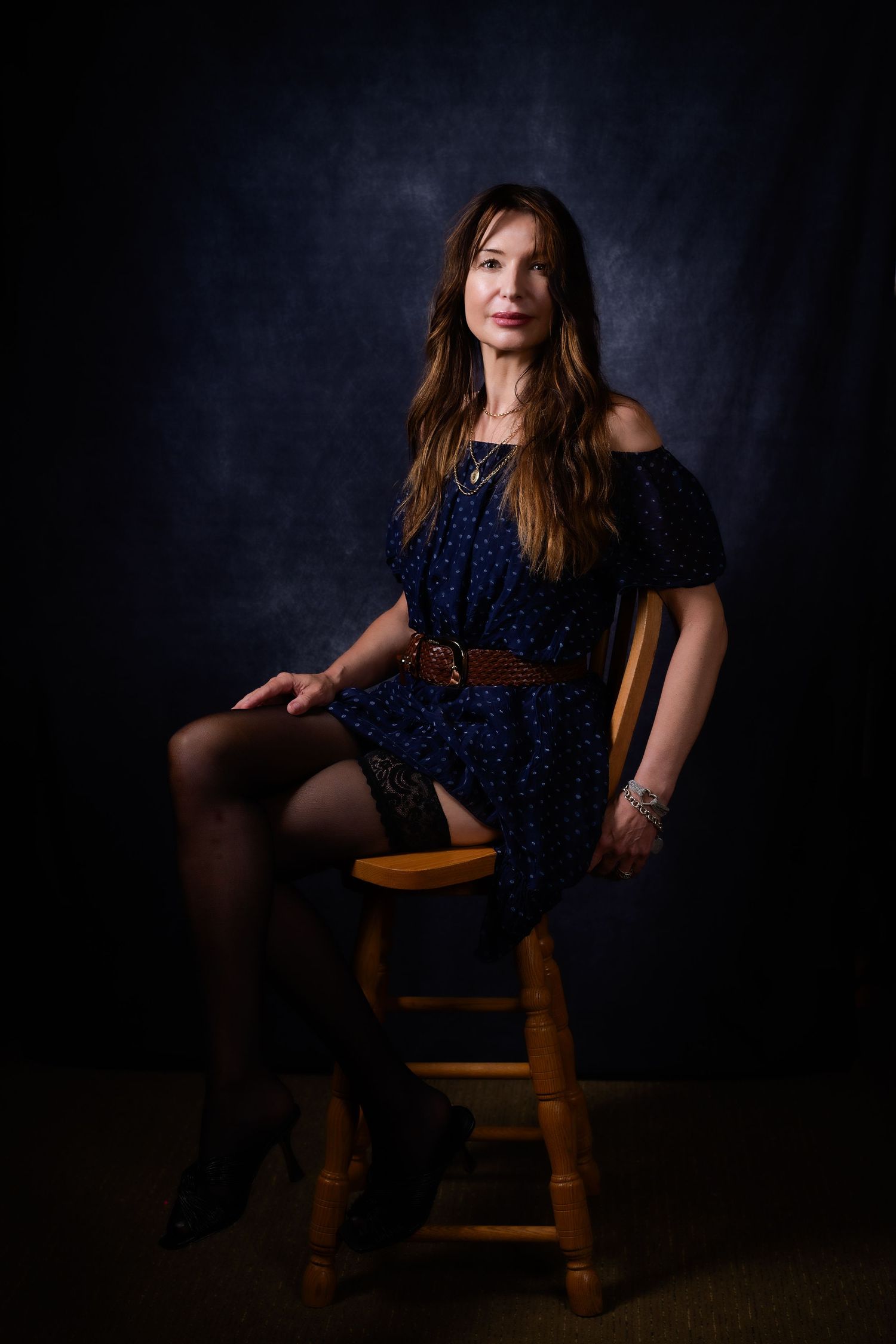 Woman in blue off-shoulder dress and tights sits on wooden stool against dark studio backdrop.